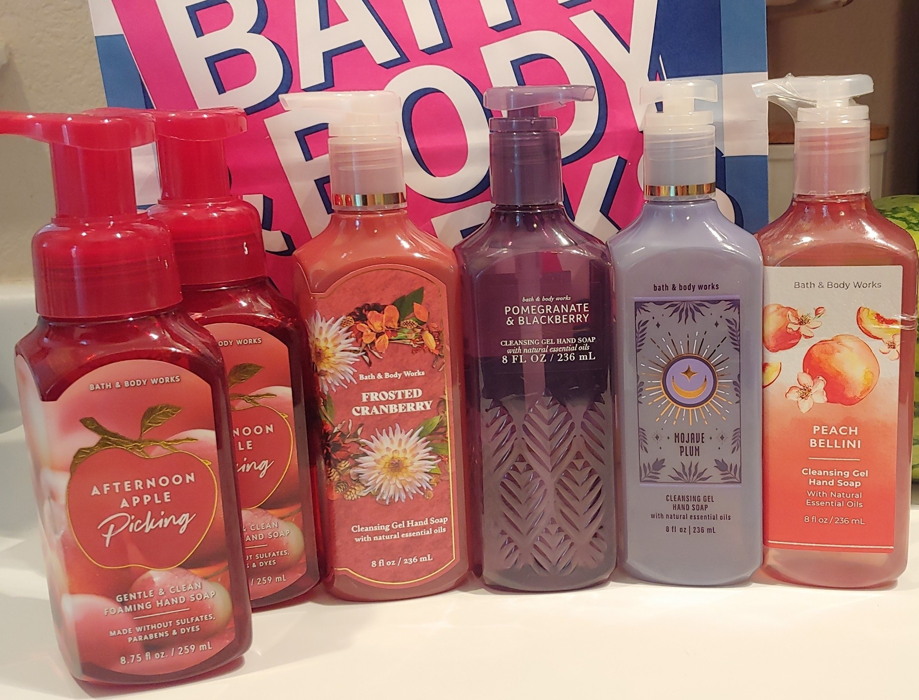 A line of six Bath and Body Works foaming and gel soap bottles in front of a Bath and Body Works bag. There are two Afternoon Apple Picking foaming soaps and one each of the four gel soaps, Frosted Cranberry, Pomegranate and Blackberry, Mojave Plum and Peach Bellini. 