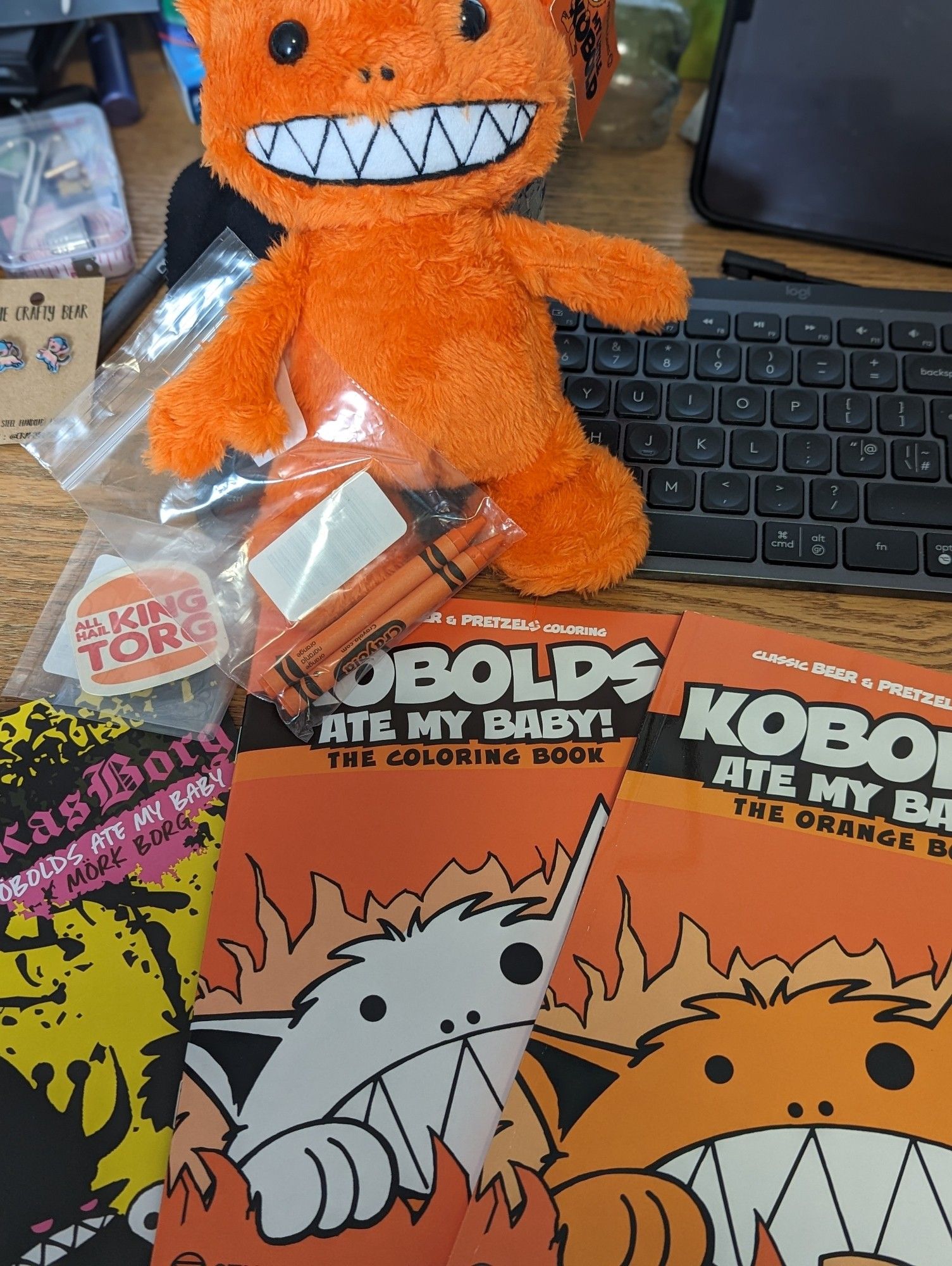 A picture of all of the loot from the backerkit crowdfunding project - Kobolds ate my baby!. There is one very cute orange kobold plushie with a wide toothy grin. A sticker shaped like a burger king icon with the words 'All Hail King Torg', a Kobolds ate my baby colouring booka Kobolds ate my baby RPG game book, a kobolds ate my baby/Mörk Borg rpb game zine thing and 2 orange Crayola crayons (for the colouring book).