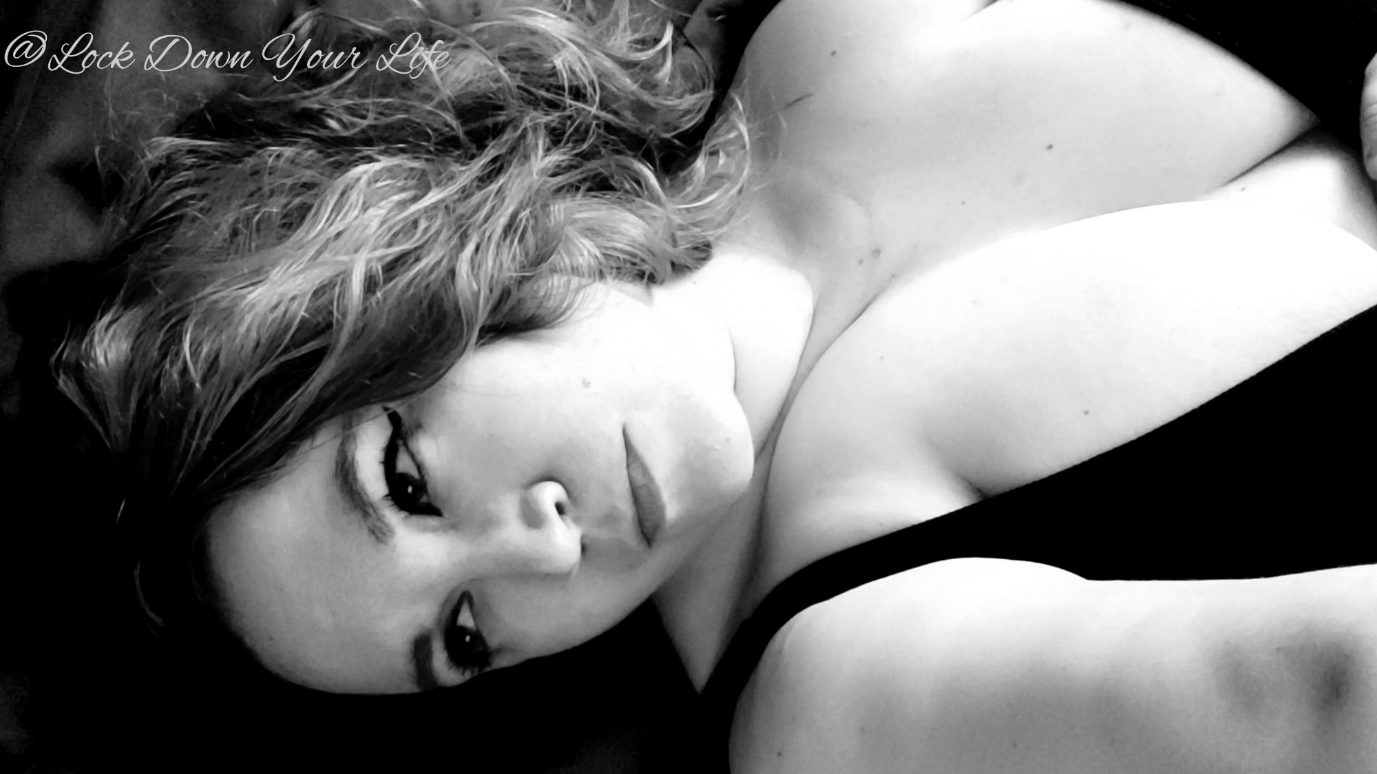 A black and white photo of LDYL wearing a black tank top and with her hair curled to one side of her head. She is lying on sheets and the photo os retro styled with winged liner and red lip. 
