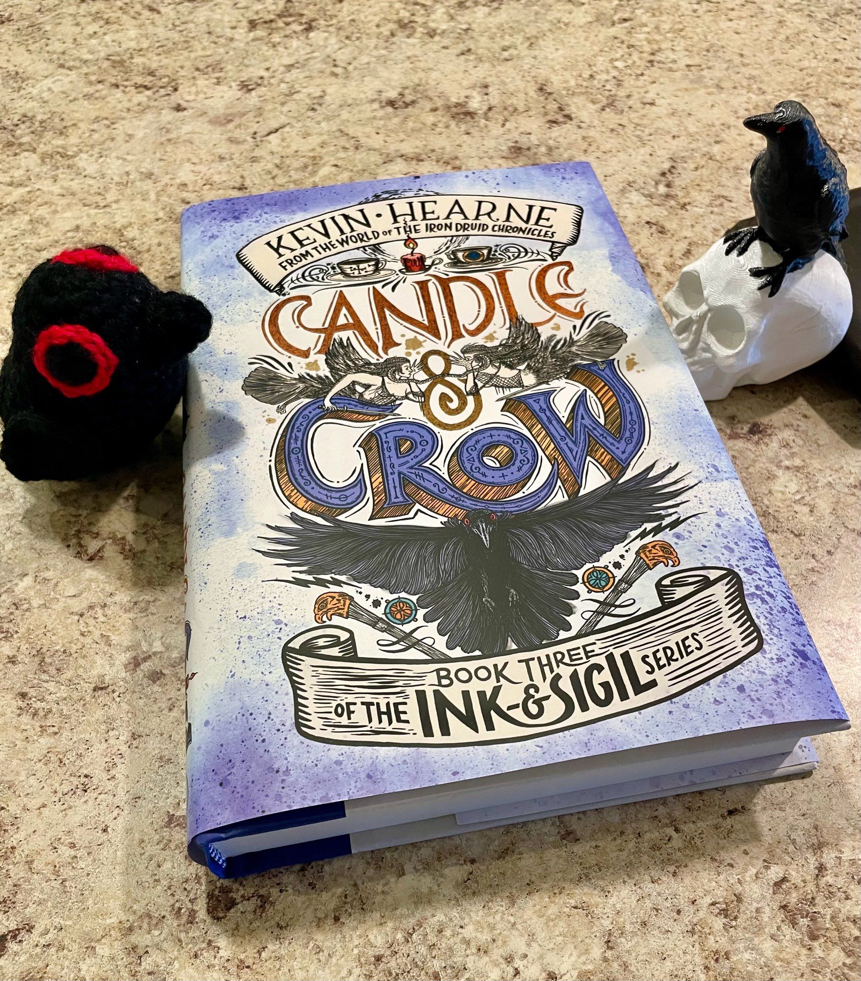 A small crocheted black crow on the left of a copy of CANDLE & CROW, plus a 3D printed crow and skull on the right 