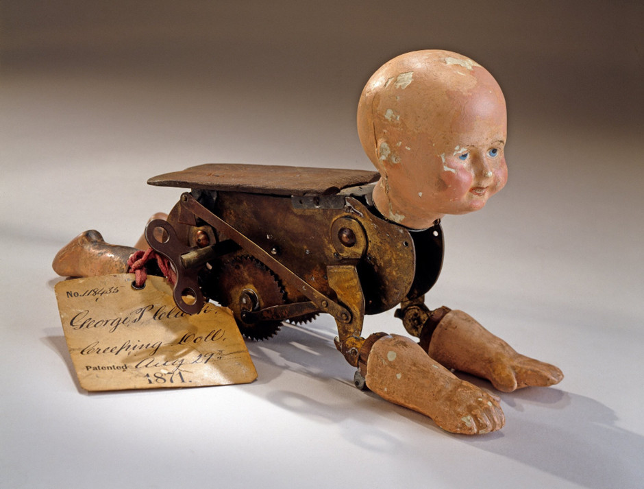 Image of a doll on all fours. Its head, two arms and two legs are made of painted plaster. The arms and legs are hinged to a brass clockwork body that moves the arms and legs in imitation of crawling, but the doll moves forward by rolling along on two toothed wheels. A flat piece of wood is attached to top of the movement.