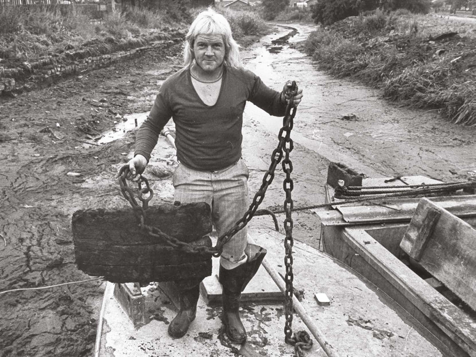 A man with a spectacular 1970s mullet, wearing big wellies and a v-neck jumper over a T-shirt, looks at camera as he stands on a boat which is itself standing on the muddy bottom of an empty canal. He is holding up an enormous wooden plug on a long chunky chain