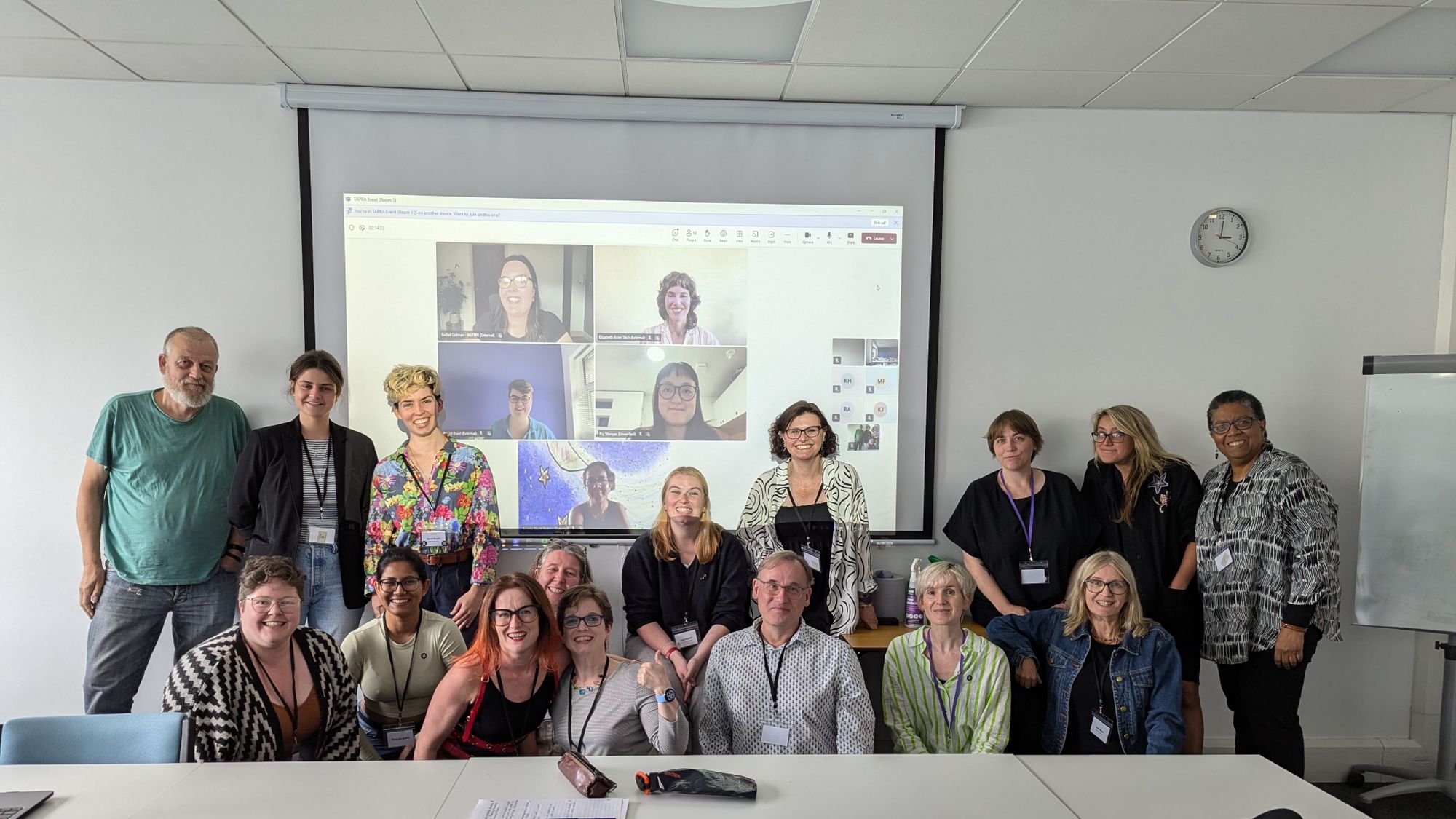 A room full of working room participants in person gathered around a screen featuring those attending virtually