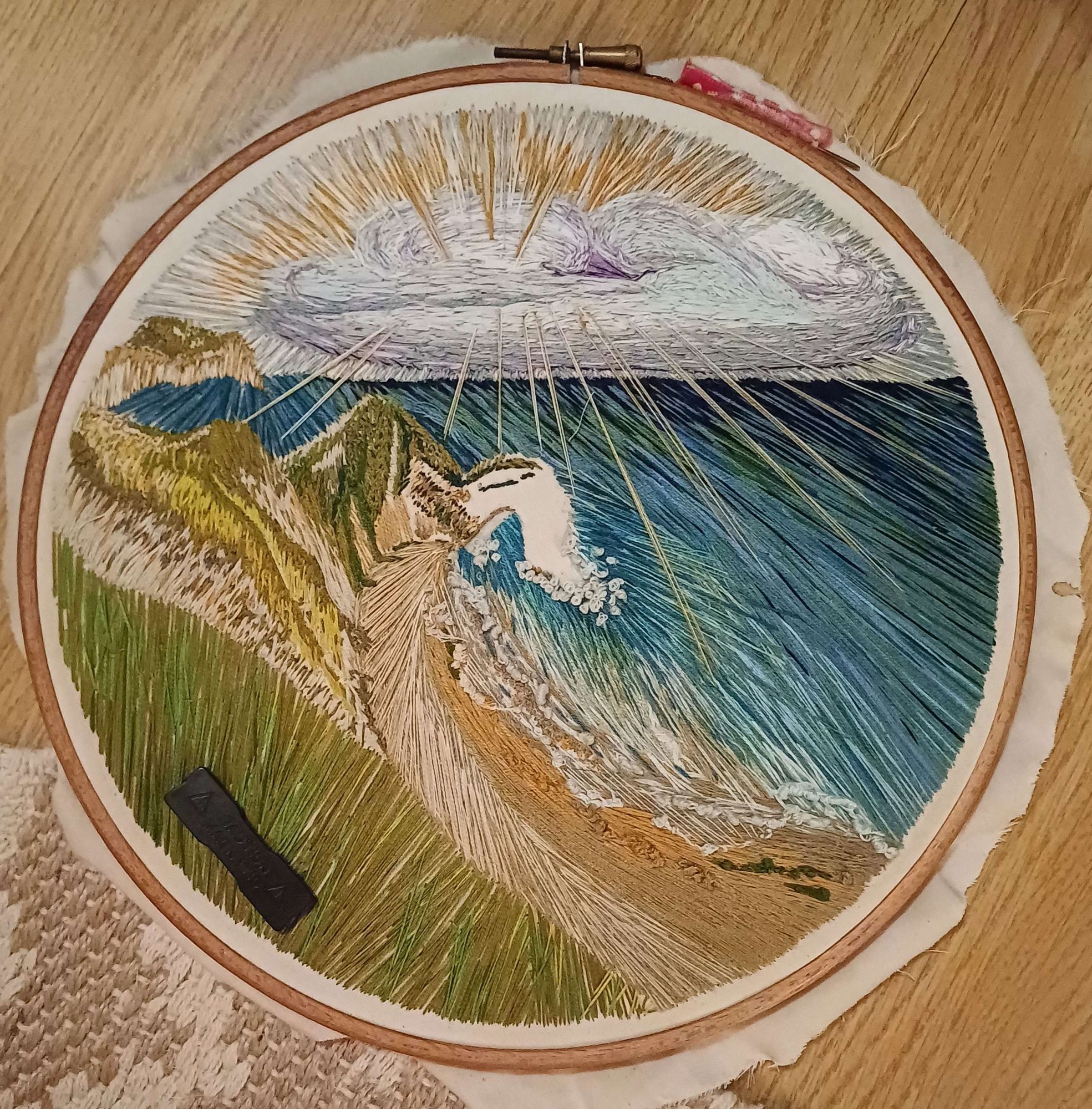 Seascape embroidery of Durdle Door in Dorset, taken from atop the cliffs looking east through the Door. Sun rays escape through a purple grey cloud across and blue sea to the foamy shore