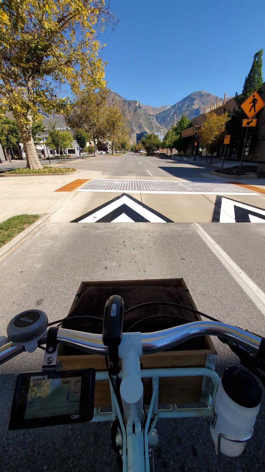 A photo taken over the handlebars of my bicycle riding down the street. Directly ahead of me across my path is a raised crosswalk with freshly painted warning chevrons. Both sides of the street have orange and yellow autumnal trees. In the distance mountains are bedecked with similar foliage.