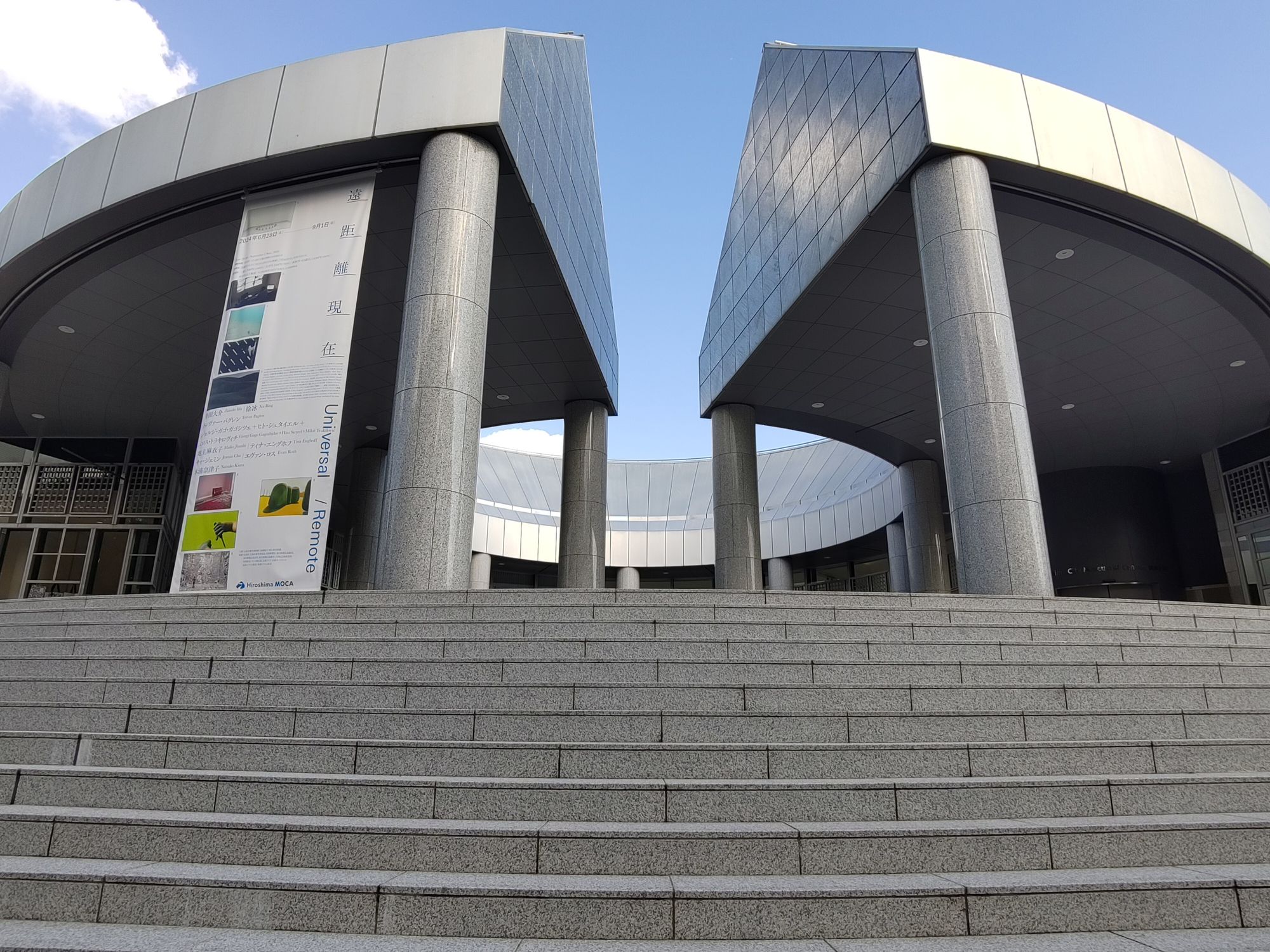 Man sieht den Eingang des Moca in Hiroshima. Der Blick geht eine grau Treppe hinauf und zeigt dann ein sehr futuristisches Gebäude in glänzenden Silber. Das Gebäude steht sehr weit oben, aber das erkennt man auf dem Bild nicht. 😃