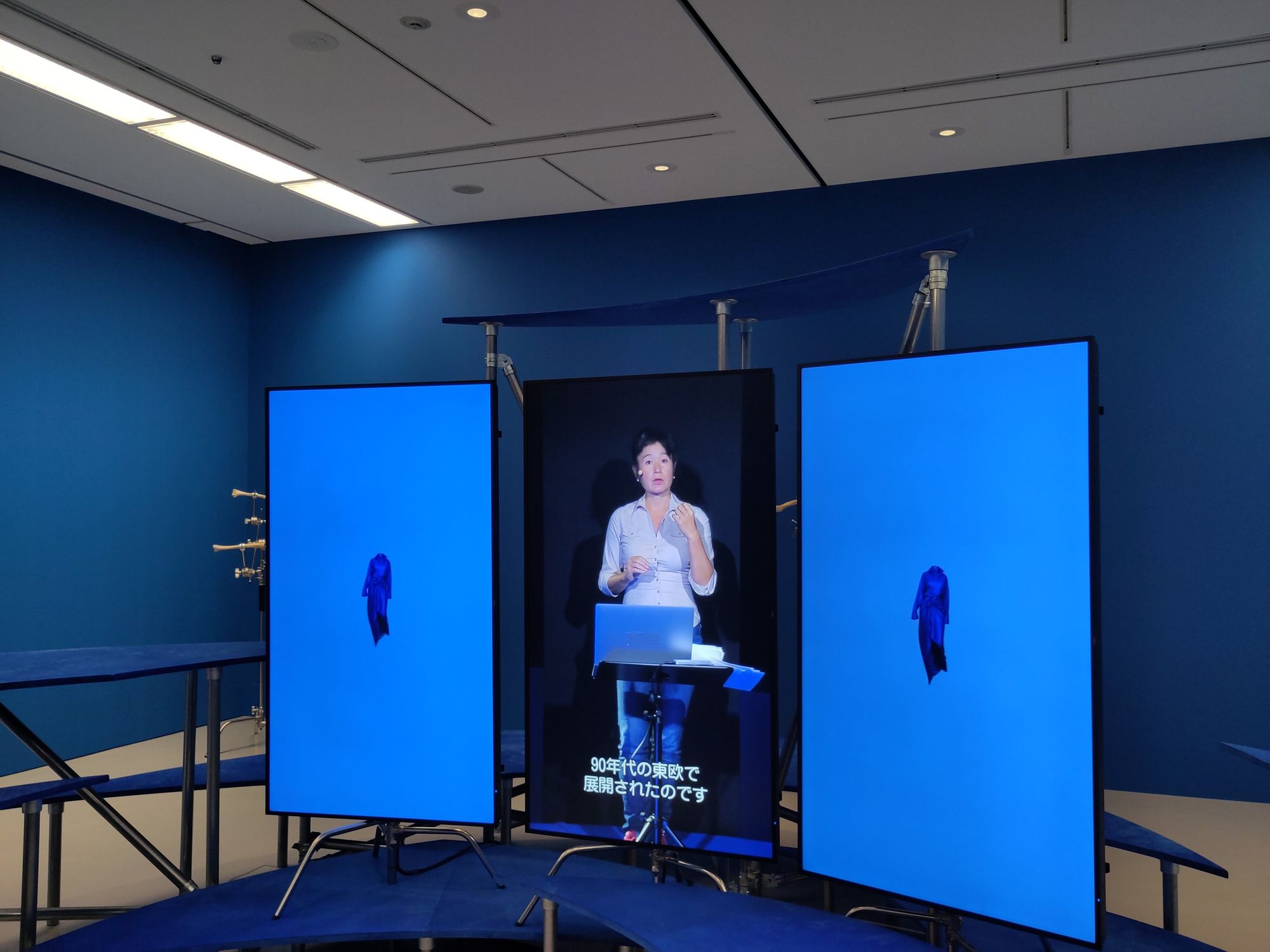 Ein Bild mit blauem Hintergrund und 3 blauen Screens. Rechts und links ist auf dem Screen ein blaues Bild zu sehen. In der Mitte sieht man Hito Steyerl hinter einem Laptop stehen und sprechen. 