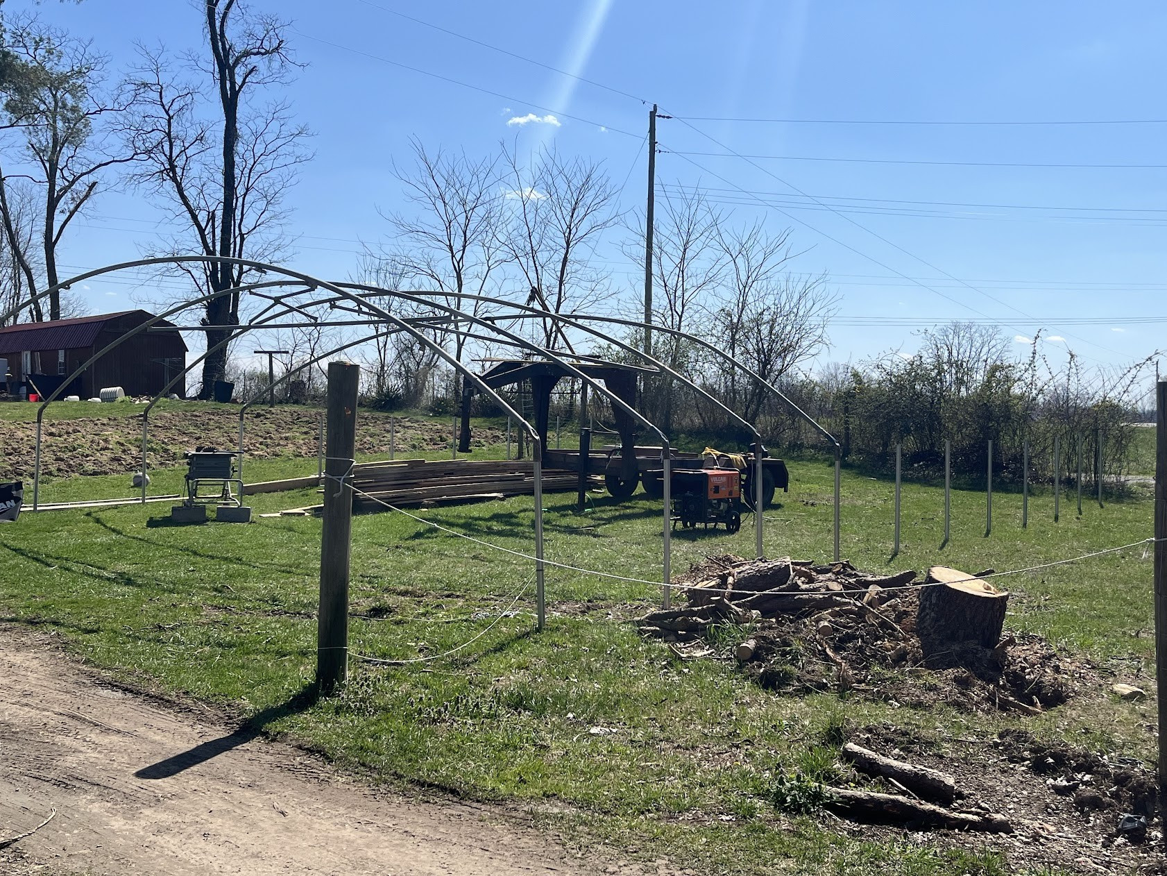 Picture of a skeleton frame of a metal greenhouse being erected. There is a gooseneck trailer that is acting as a mobile gin pole (a device used to lift hoops into position and keep them from twisting as they are fitted over pipes driven into the ground). There is a welder/generator in the picture because when the greenhouse was used by the previous owners, they made modifications that need to be fixed for our purposes