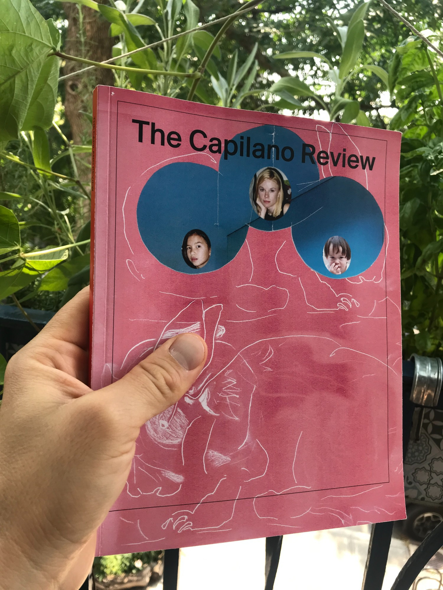 White left hand holding the latest issue of The Capilano Review against a leafy balcony, displaying cover art by Marisa Kriangwiwat Holmes.