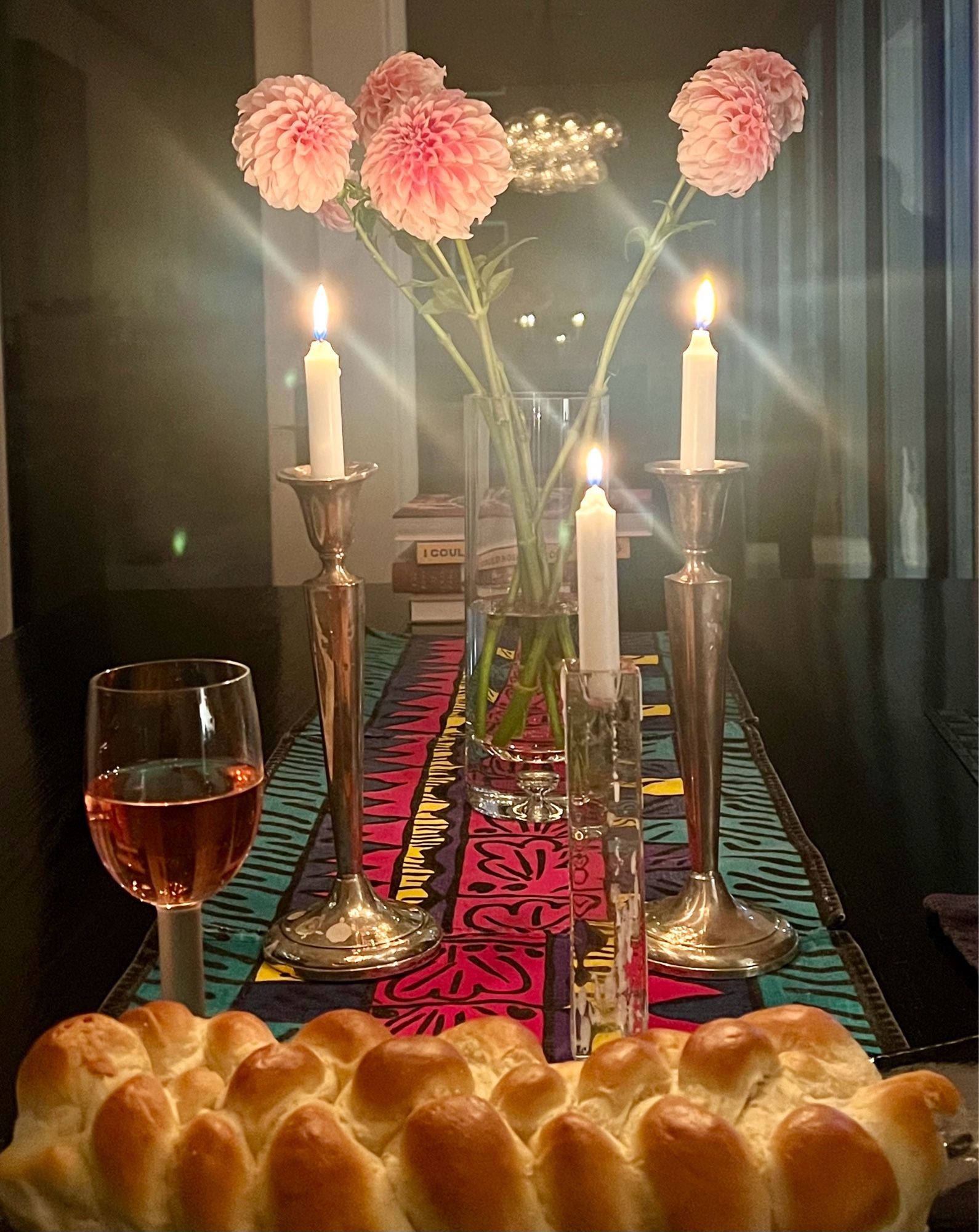 A Shabbat table in front of a window at night. Vase of dahlia zinnias; a tall pair of lit candles; a third one for hope and the hostages; a wine glass; challah.