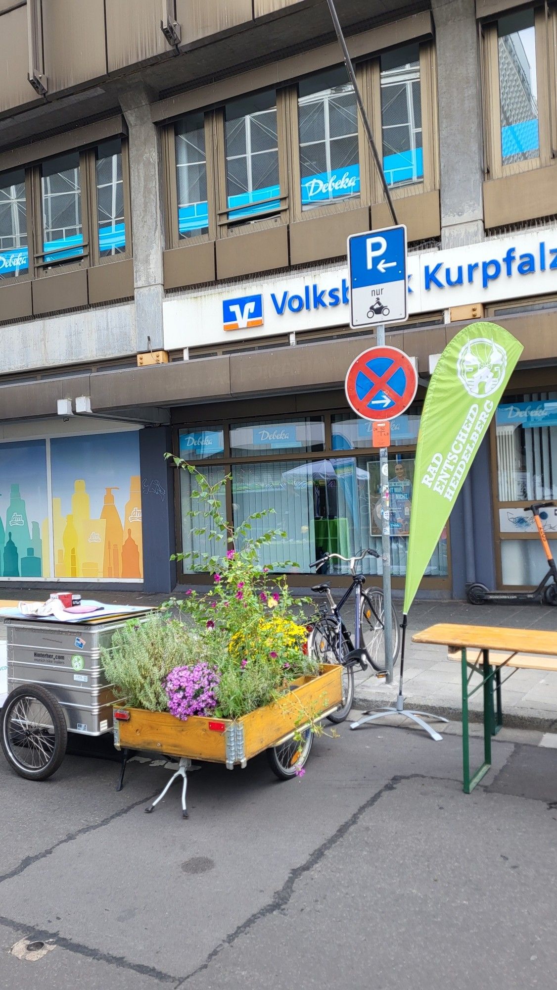 Ein Hinterher-Anhänger, ein bepflanzter Fahrradanänger mit bunten Farben und eine Biertischgarnitur am Fahrbahnrand.