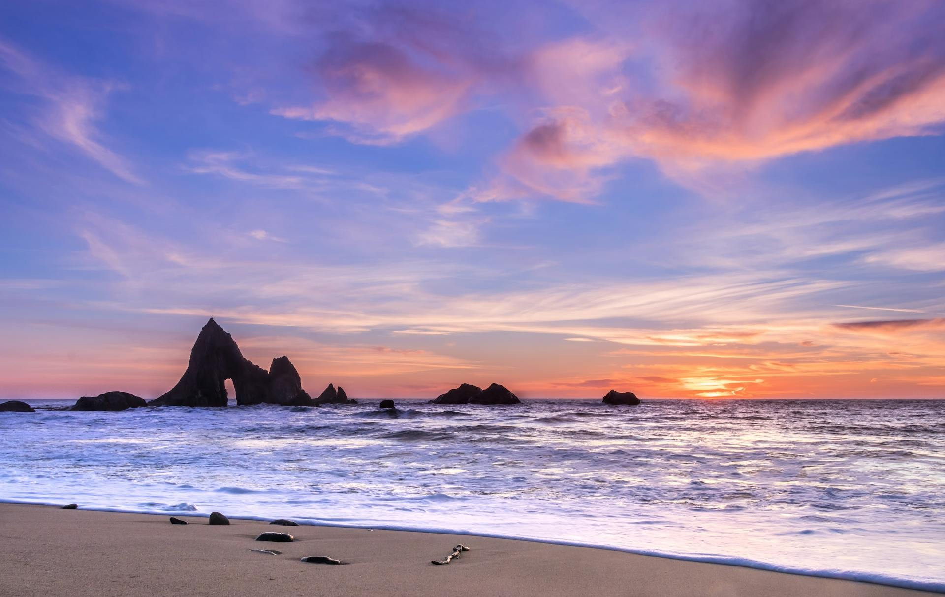 The sun sets on the horizon over the ocean with rock formations in the water near the shoreline.