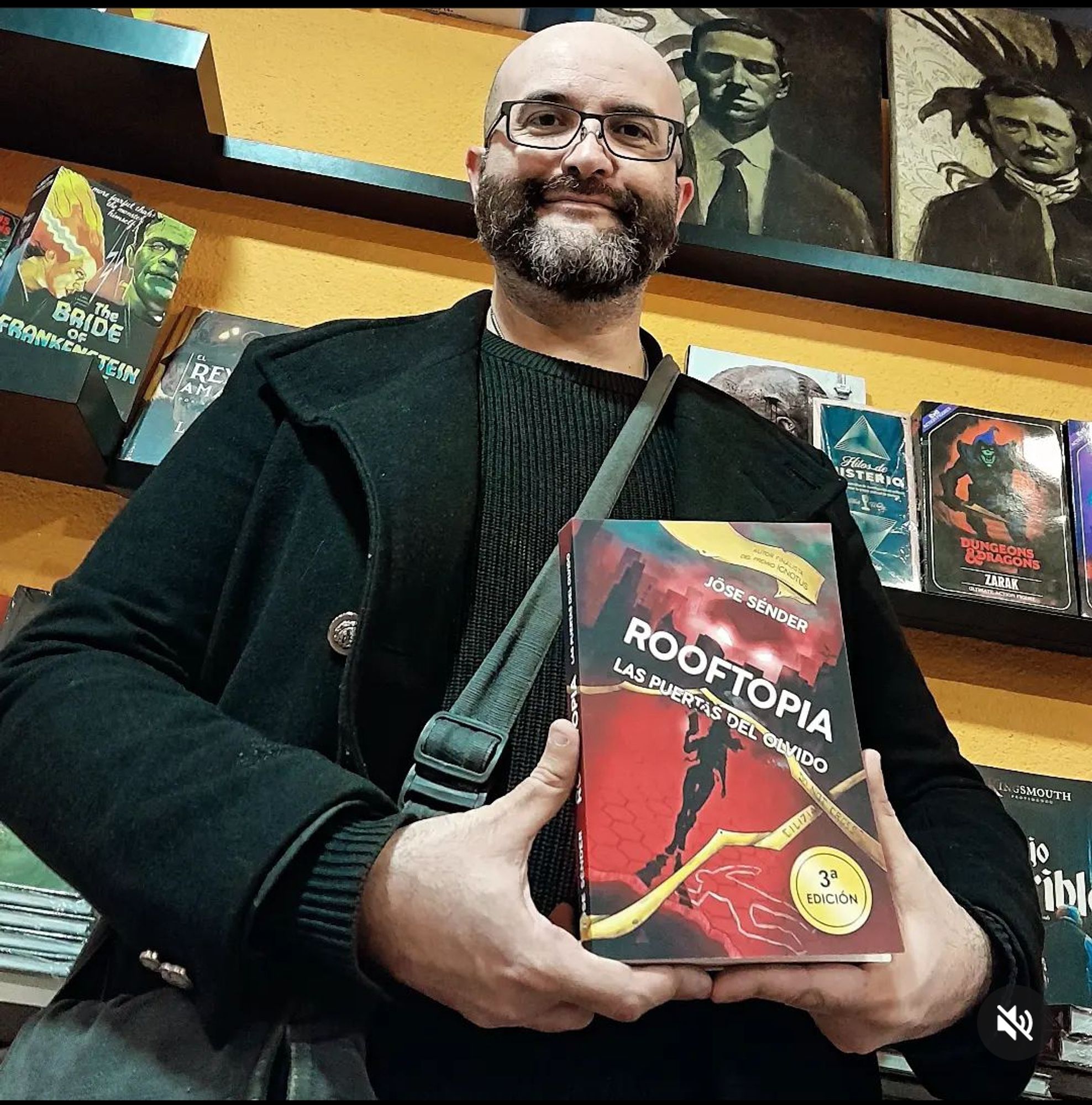 El escritor Jose Sénder en la librería Landròmina, sosteniendo una copia de su novela Rooftopia, delante de sendos retratos de H.P. Lovecraft y Edgar Alan Poe.