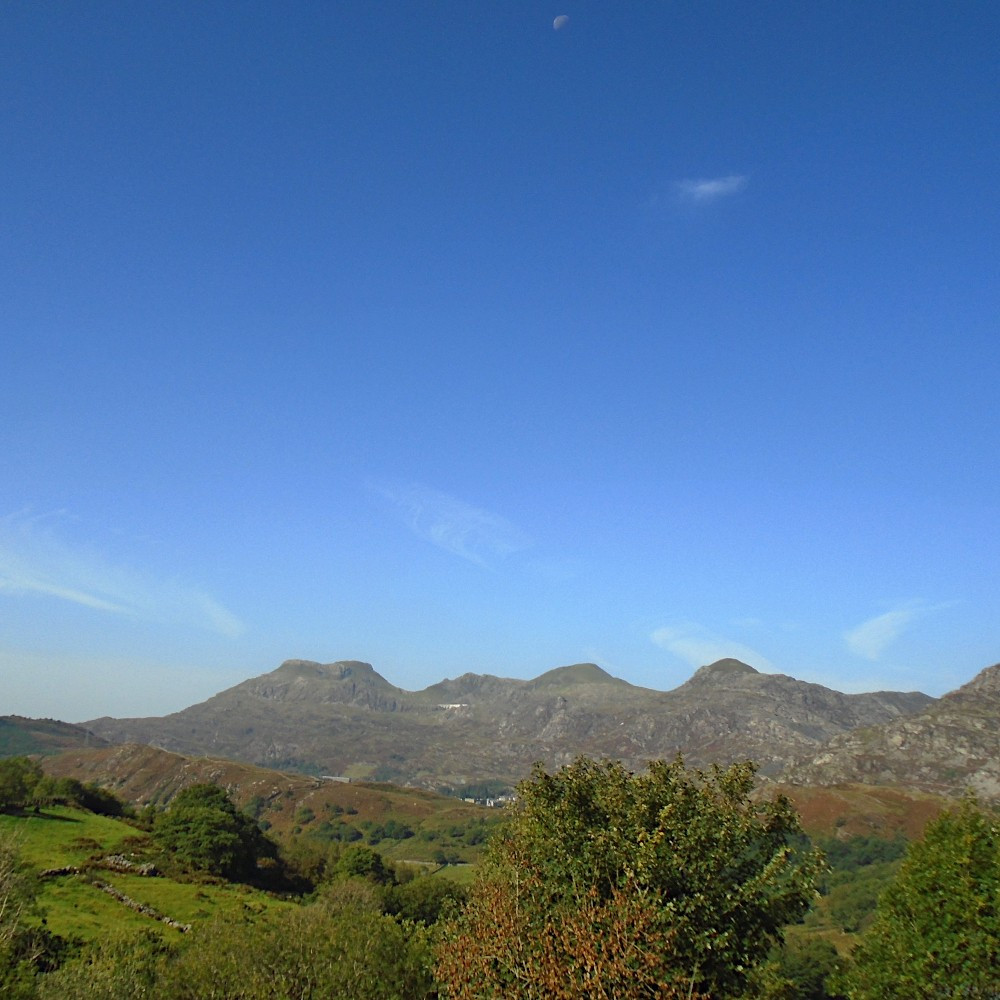 a few wispy clouds in an otherwise blue sky, with the moon still visible at the top of the frame, high above a brightly lit series of valleys and slightly hazy, distant mountains