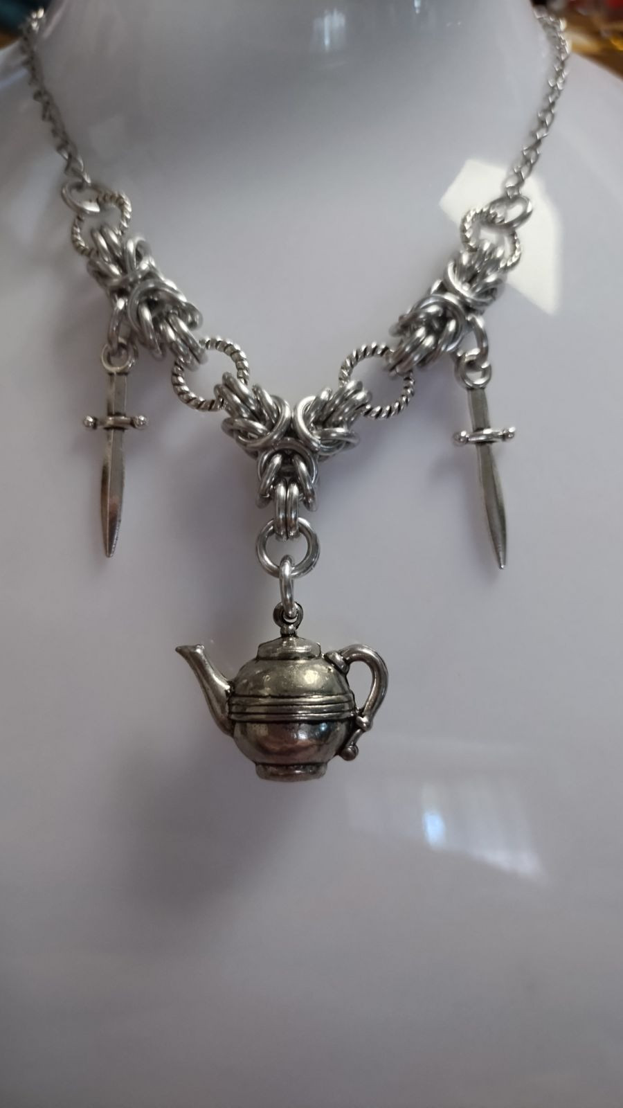 chainmail necklace with a teapot charm hanging down from the center and a dagger charm on either side on a clear and white plastic necklace display