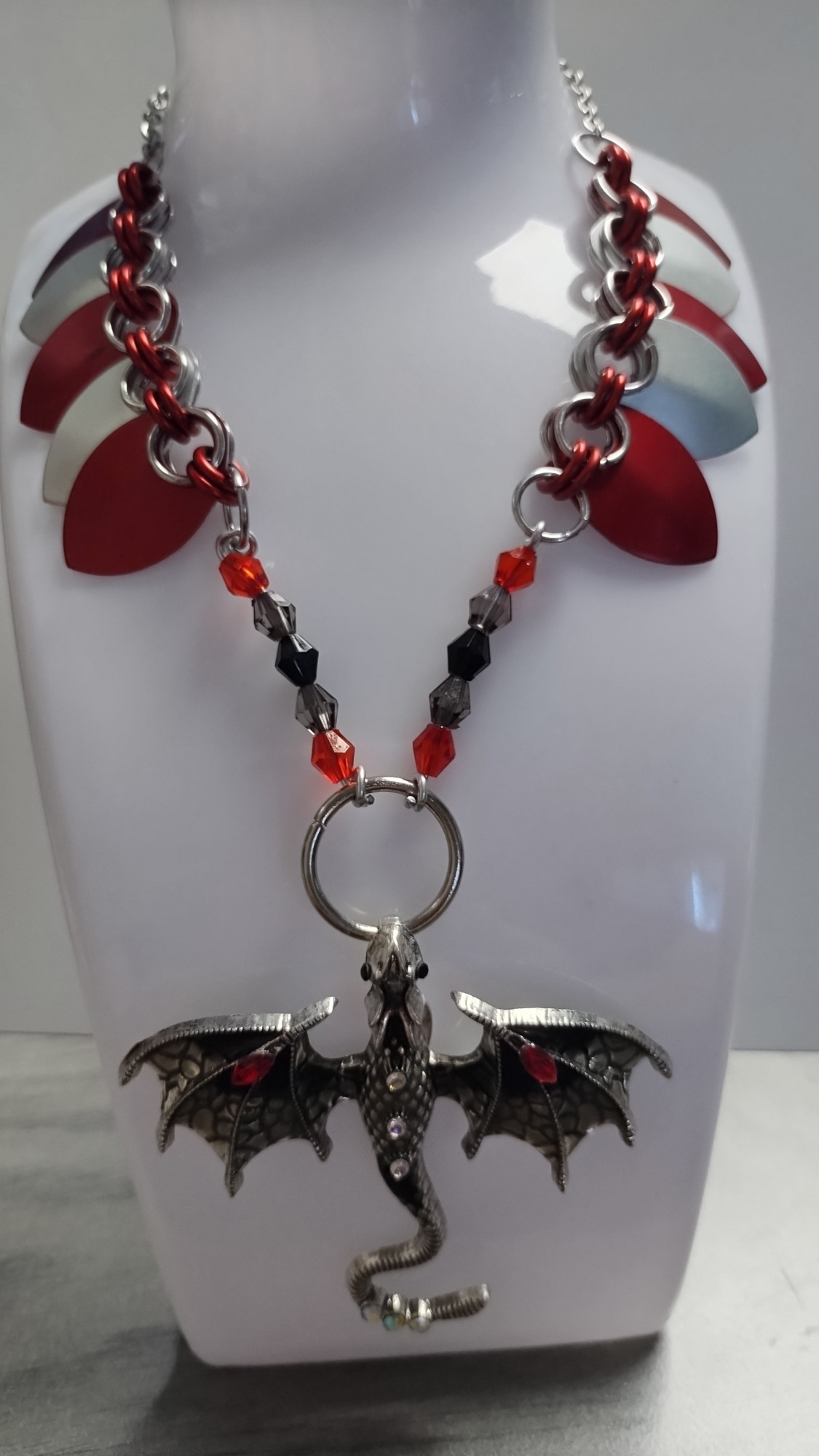 Photo of a chainmail and scalemail necklace on a white plastic holder in front of a glossy white background. The holder is sitting on a grey wood desk. A red, black, and silver dragon pendant hangs beneath red and silver scales with red and black bead elements.