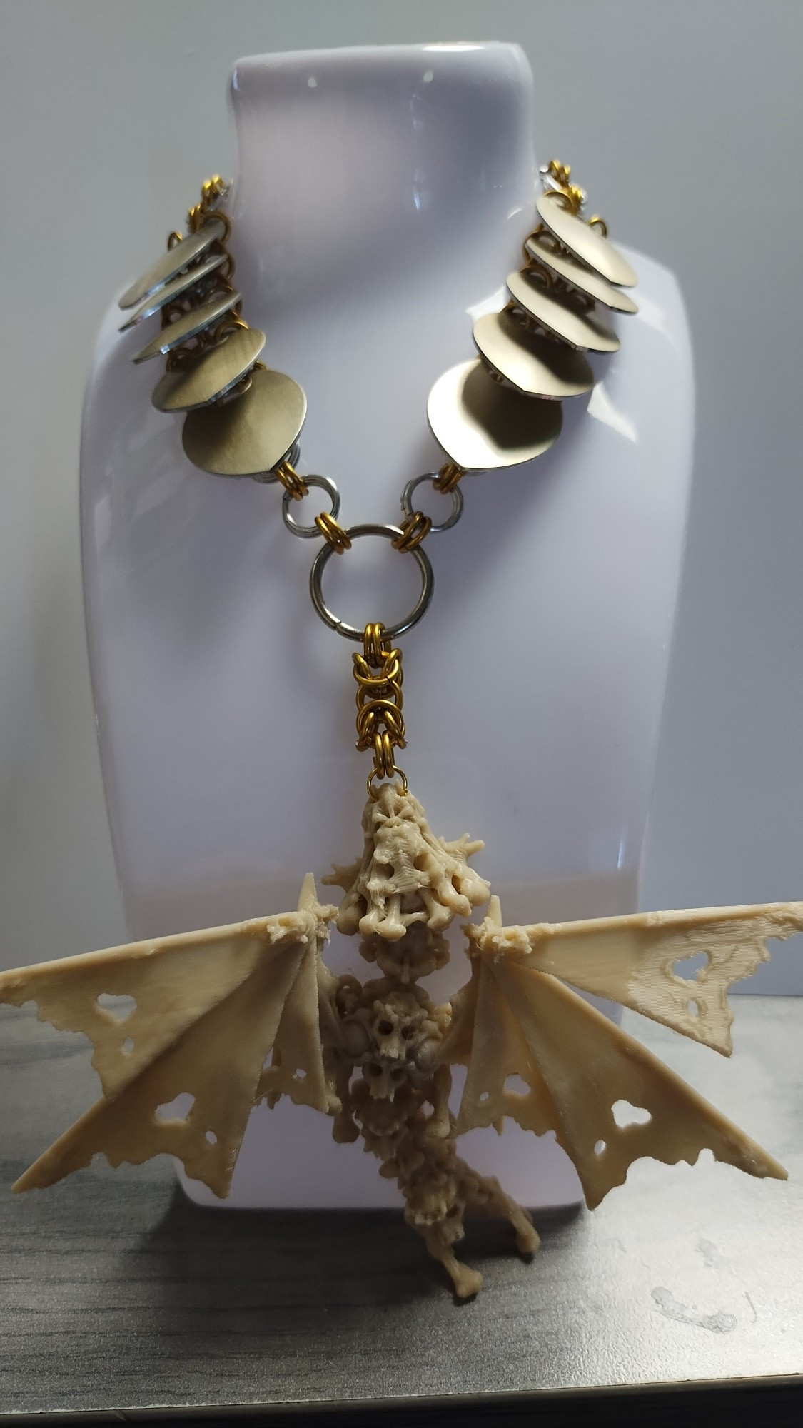 Photo of a chainmail and scalemail necklace on a white plastic holder in front of a glossy white background. The holder is sitting on a grey wood desk.  The dragon pendant is 3d printed in bone white plastic and the scales are champagne gold