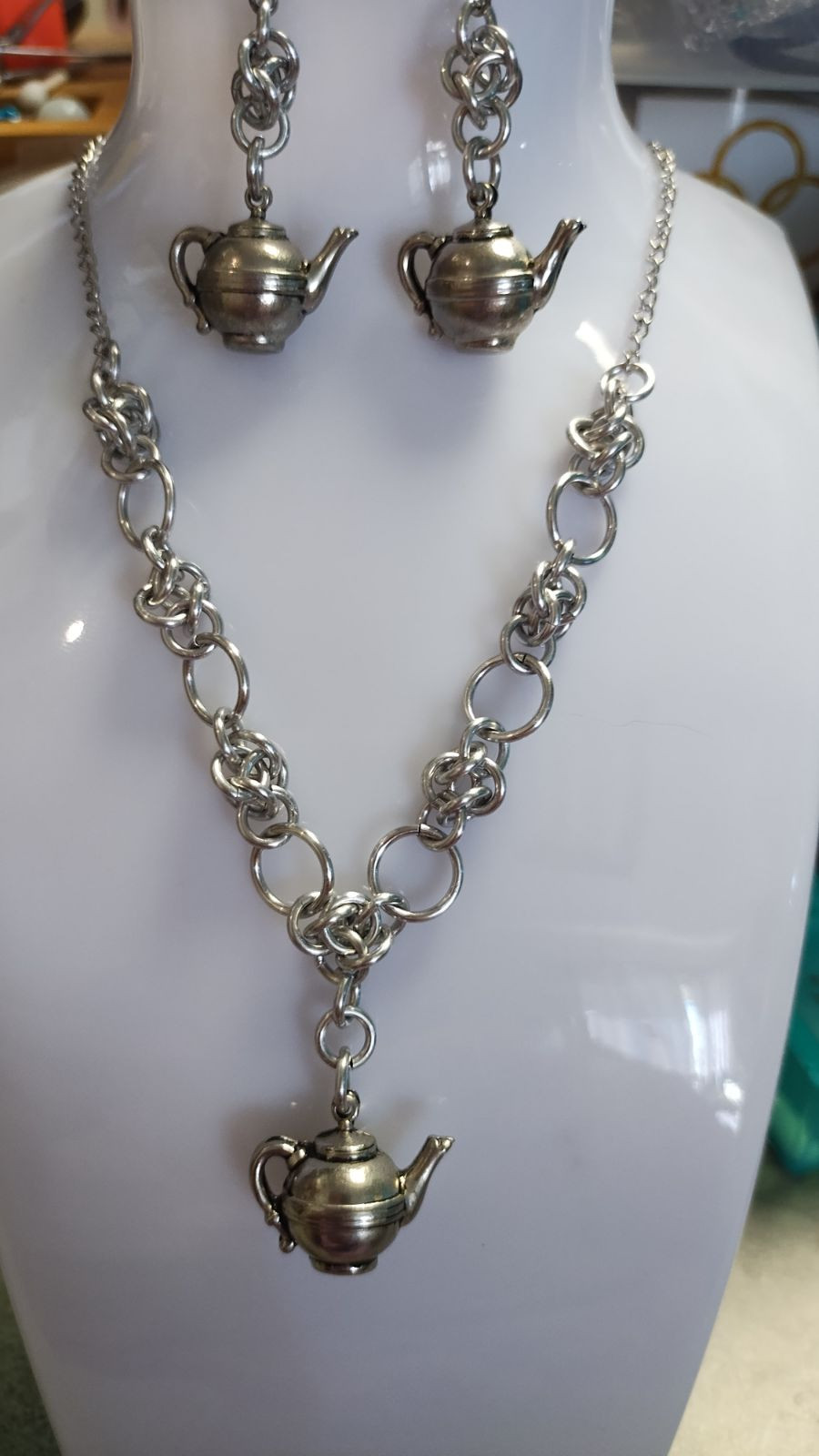 chainmail necklace with a teapot charm hanging down from the center on a clear and white plastic necklace display. matching earrings hang from the top