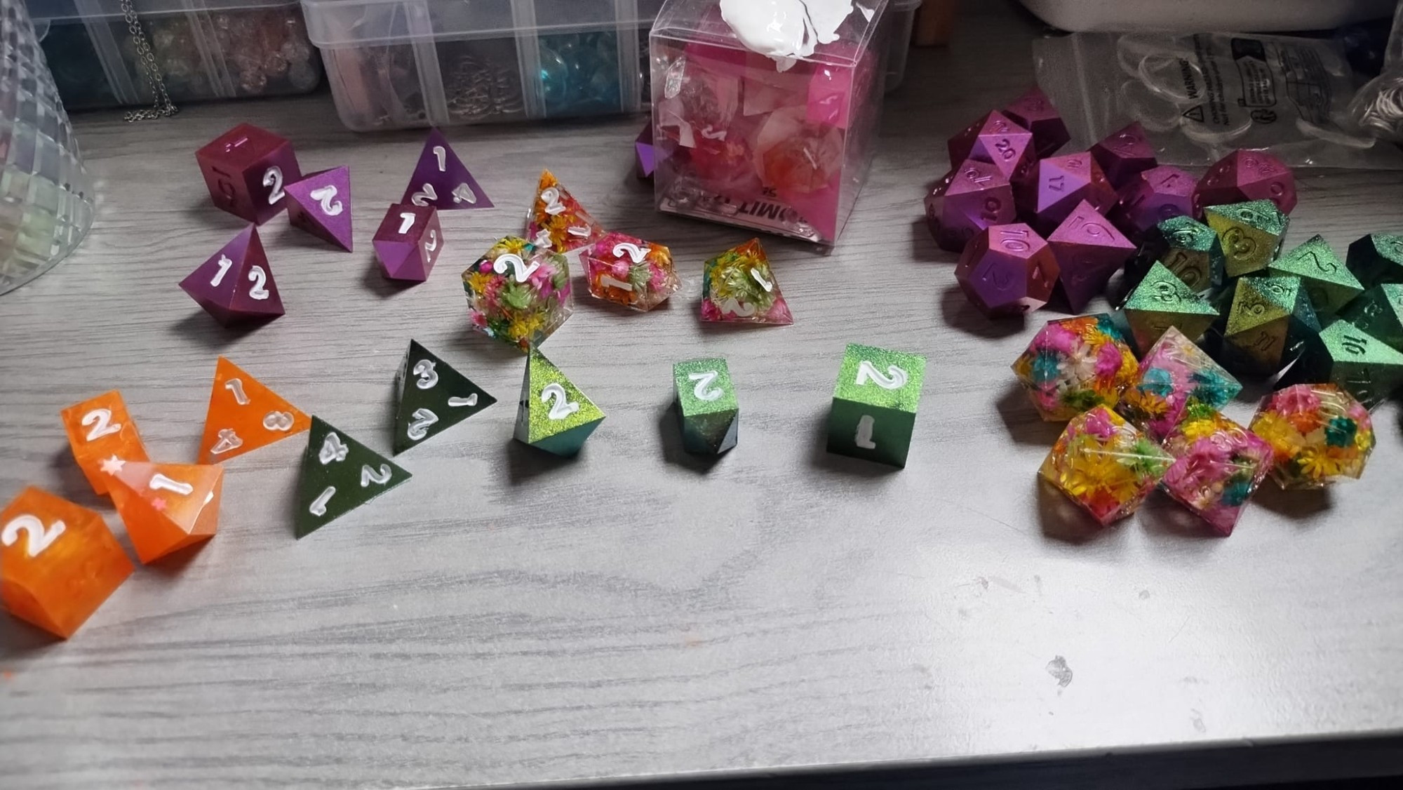 photo of a grey wooden desktop with assorted dice spread across it, some inked in white and others yet unpainted. The dice are in groups by color of orange, green metallic, magenta metallic, or bright floral filled dice