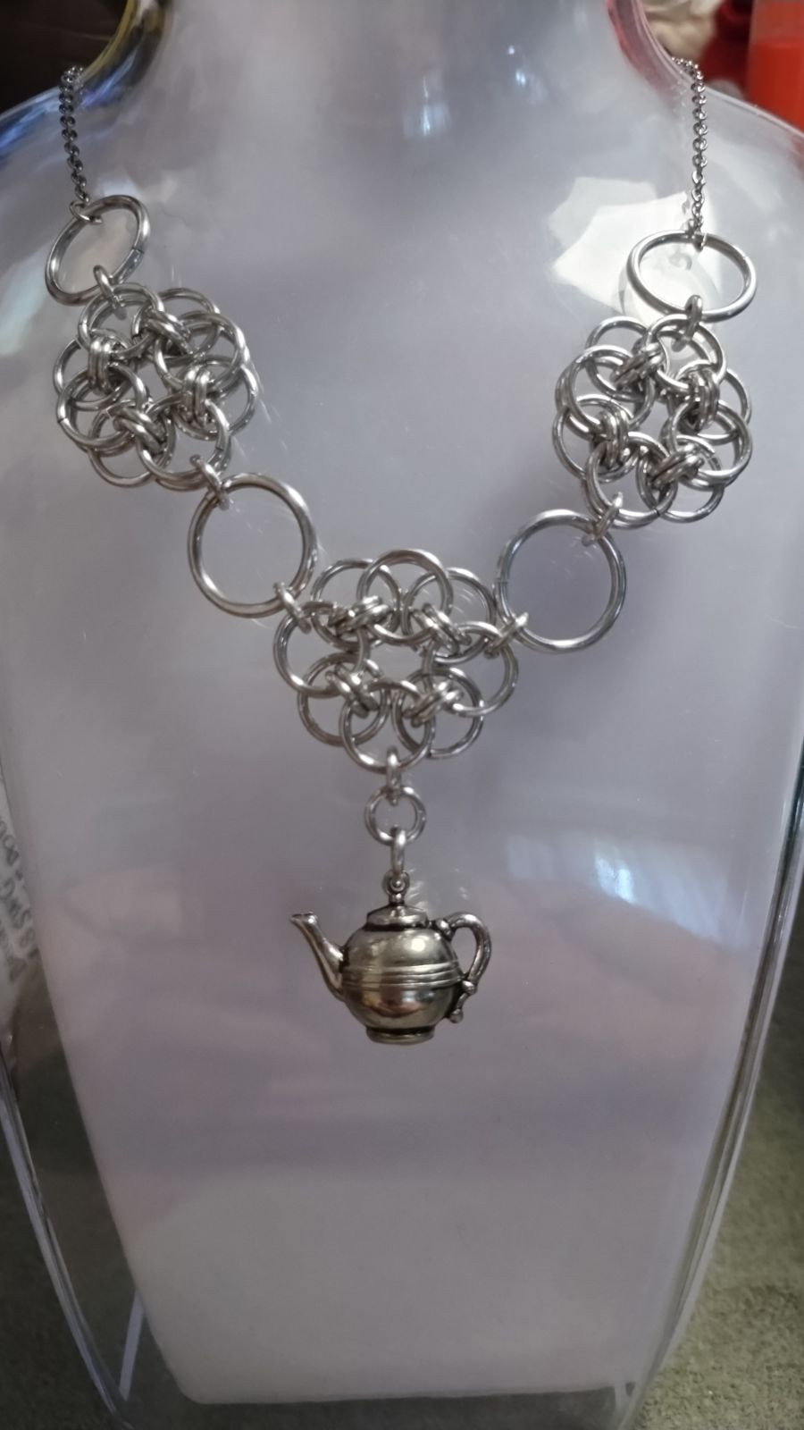 chainmail necklace of circular flower shapes with a teapot charm hanging down from the center on a clear and white plastic necklace display