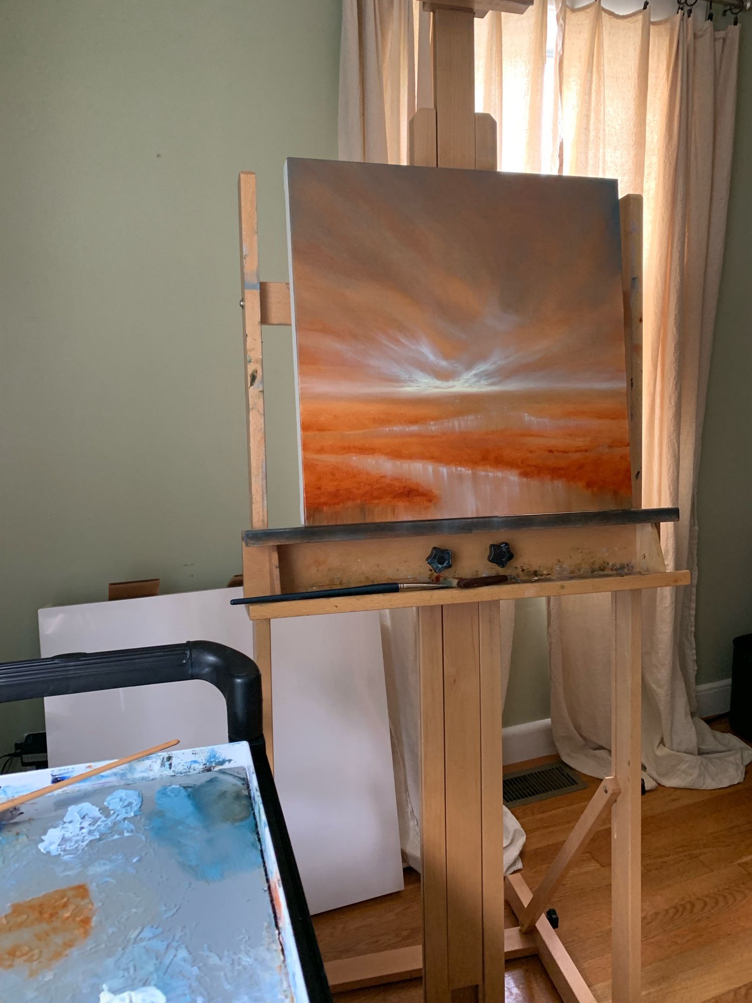 Photo of a coastal marsh oil painting in progress by Tisha Mark. The painting rests on an easel and has an orange toned sunrise sky over a coastal marsh. There is a paintbrush and a palette knife resting on the shelf below the painting. A paint palette and another paintbrush are partially visible on the left. There are canvases resting against the wall behind the easel.