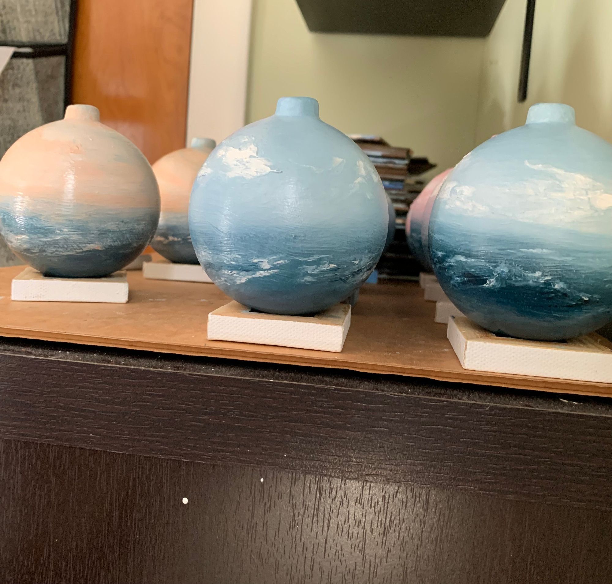 Photo of several hand-painted ornaments in progress, painted by Tisha Mark. Some have an orange-toned sky, some are all blues, and some have a pink-toned sky. They are resting on upside-down tiny canvases to keep them from rolling off the shelf.