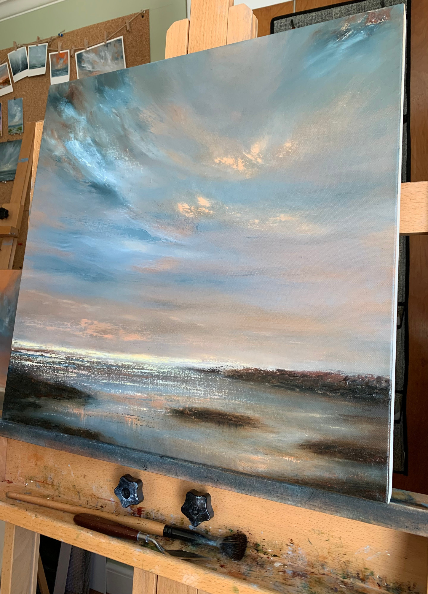 Photo of an oil painting in progress, on an easel in Tisha Mark's studio. This is a painting of a seascape at sunset, with dramatic blue cloud formations in an orange-toned sky, with flecks of orange light and a band of yellow sunlight on the horizon. Colors from the sky are reflected in the water along the rocky coast. A paintbrush and a palette knife are resting on the easel, below the painting. In the background behind the easel, some small paintings are visible on a cork board on a wall of the studio.