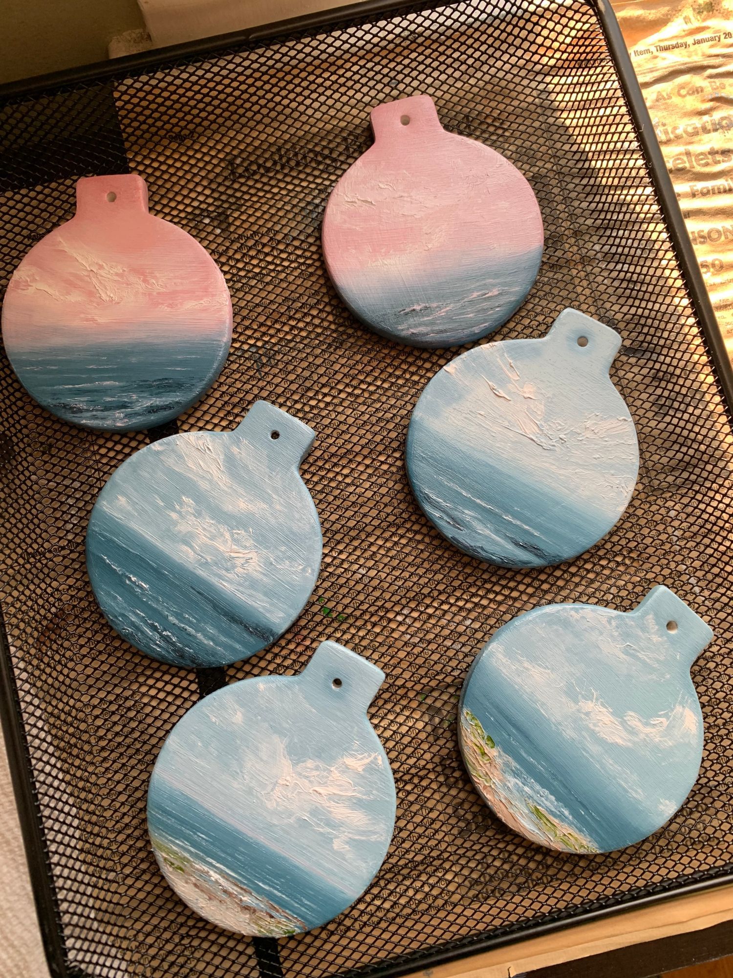 Photo of 6 ornaments, hand-painted with seascapes by Tisha Mark. The top 2 have a pink-toned sky. The middle 2 are blues and whites. the bottom 2 have beaches. They are resting on a drying rack.