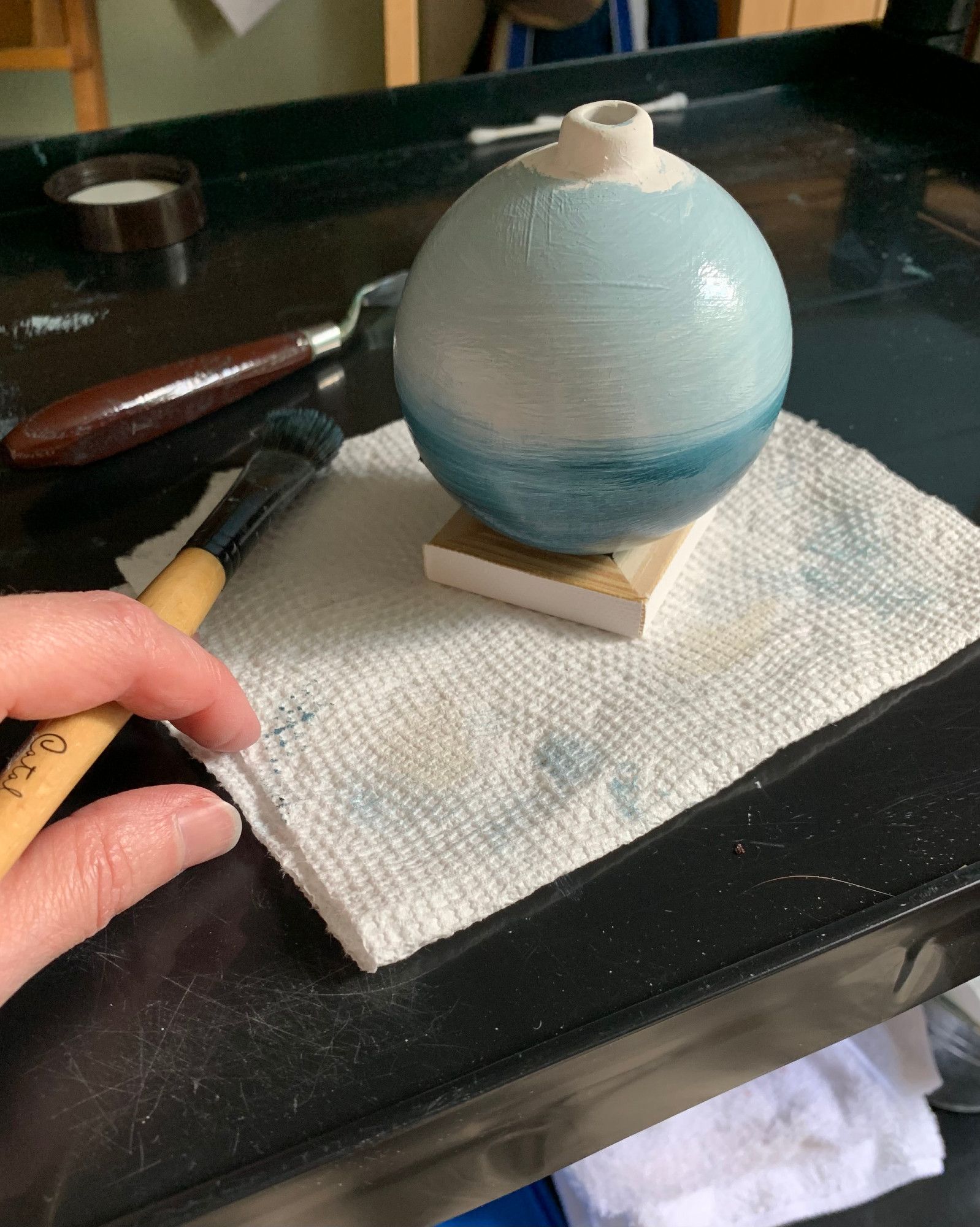 Photo of my hand holding a paint brush next to a spherical ornament that I'm painting with oil paint. The painting is in the early stages of a seascape with various shades of blue. The ornament is resting on an upside-down tiny canvas on a paper towel. In the background is a palette knife and some cotton swabs that are also used in the painting process.