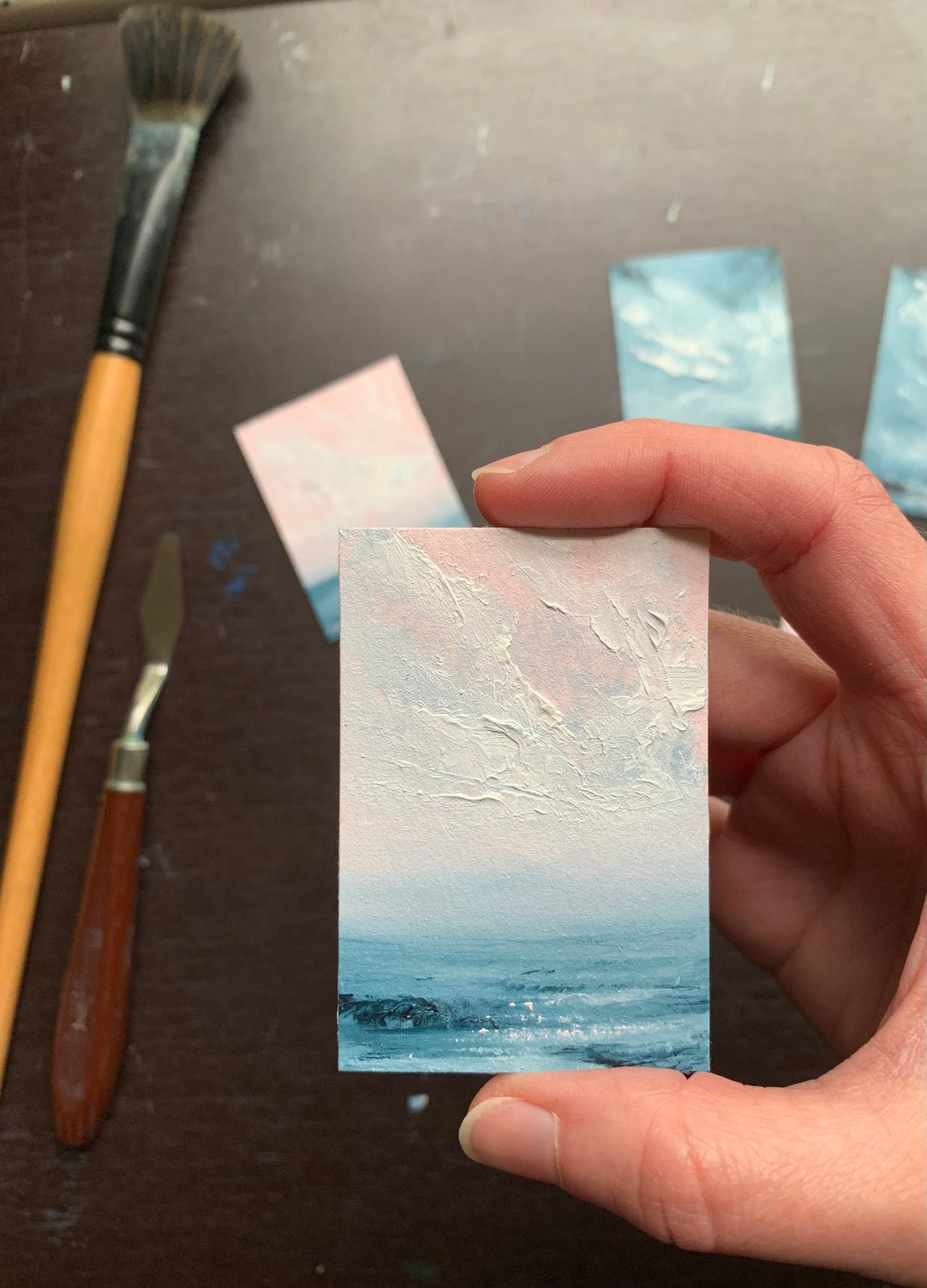 Photo of a hand holding a tiny seascape oil painting by Tisha Mark, Fine Art Card No. 2414, oil on Arches oil paper, 3"x2" (2024). The seascape is vertically oriented and is a seascape with textured cloud formations in a pink-toned sky over a deep blue sea with a rocky coast. A paintbrush, palette knife, and a few other tiny paintings are visible in the background.