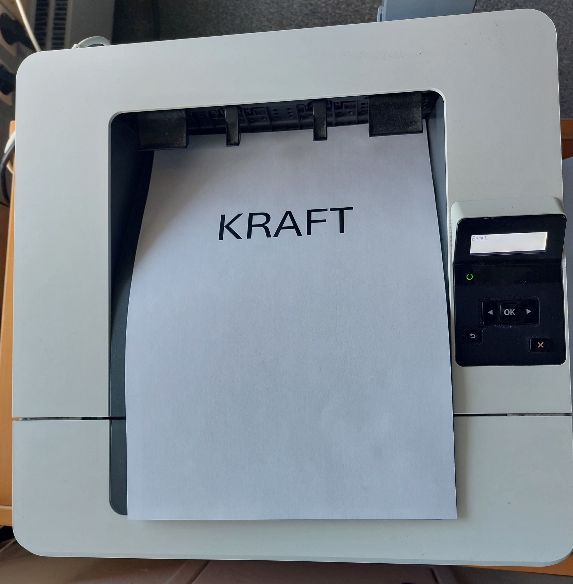 Foto eines klassischen Office Druckers. Es liegt ein frisch gedrucktes, weißes Blatt darin. Es steht nur ein Wort darauf: KRAFT