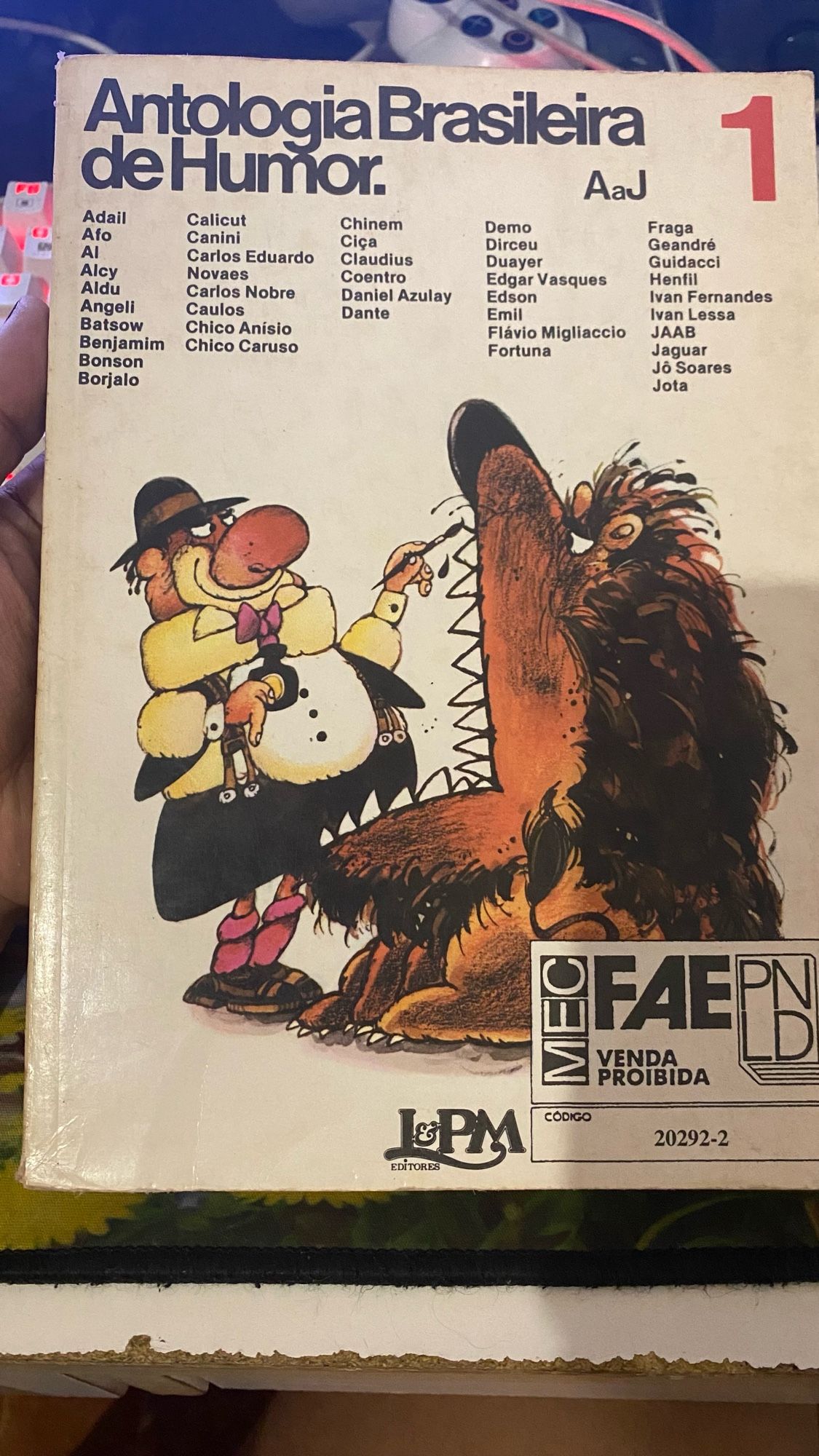 Capa de um livro chamado Antologia Brasileira de Humor. AaJ 1, com diversos nomes grafados e a ilustração de um palhaço gordo desenhando os dentes de um leão, que está com a boca bem aberta e a expressão zangada. Na capa há o selo de venda proibida do MEC.