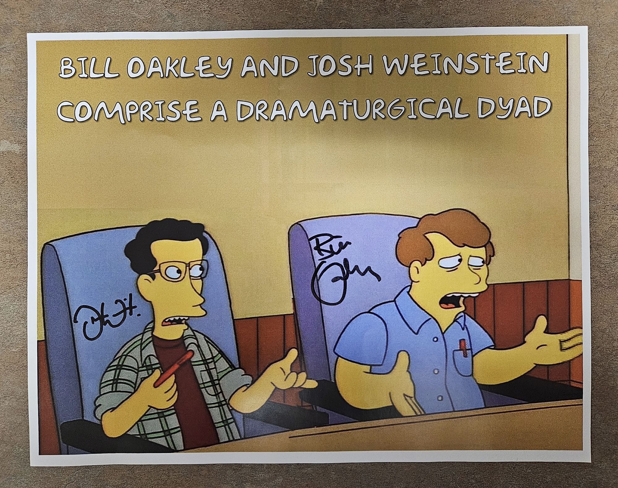 An image of Oakley & Weinstein from their appearance in The Simpsons episode "The Itchy & Scratchy & Poochie Show", sitting at a table in a writer's room, with overlaid text reading: "Bill Oakley and Josh Weinstein comprise a dramaturgical dyad."