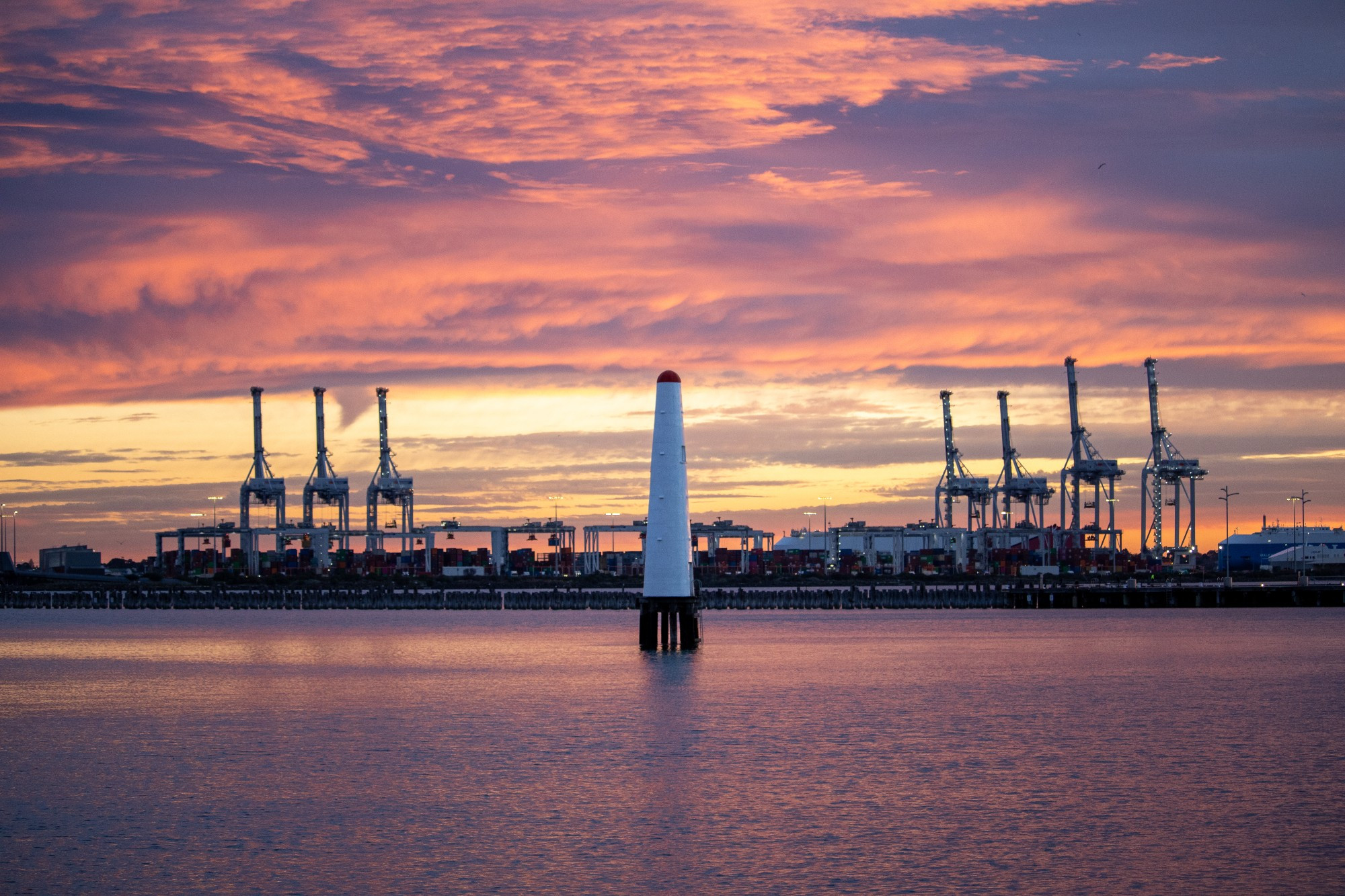 Photo of a lighthouse in the foreground between large cranes in the background at sunset