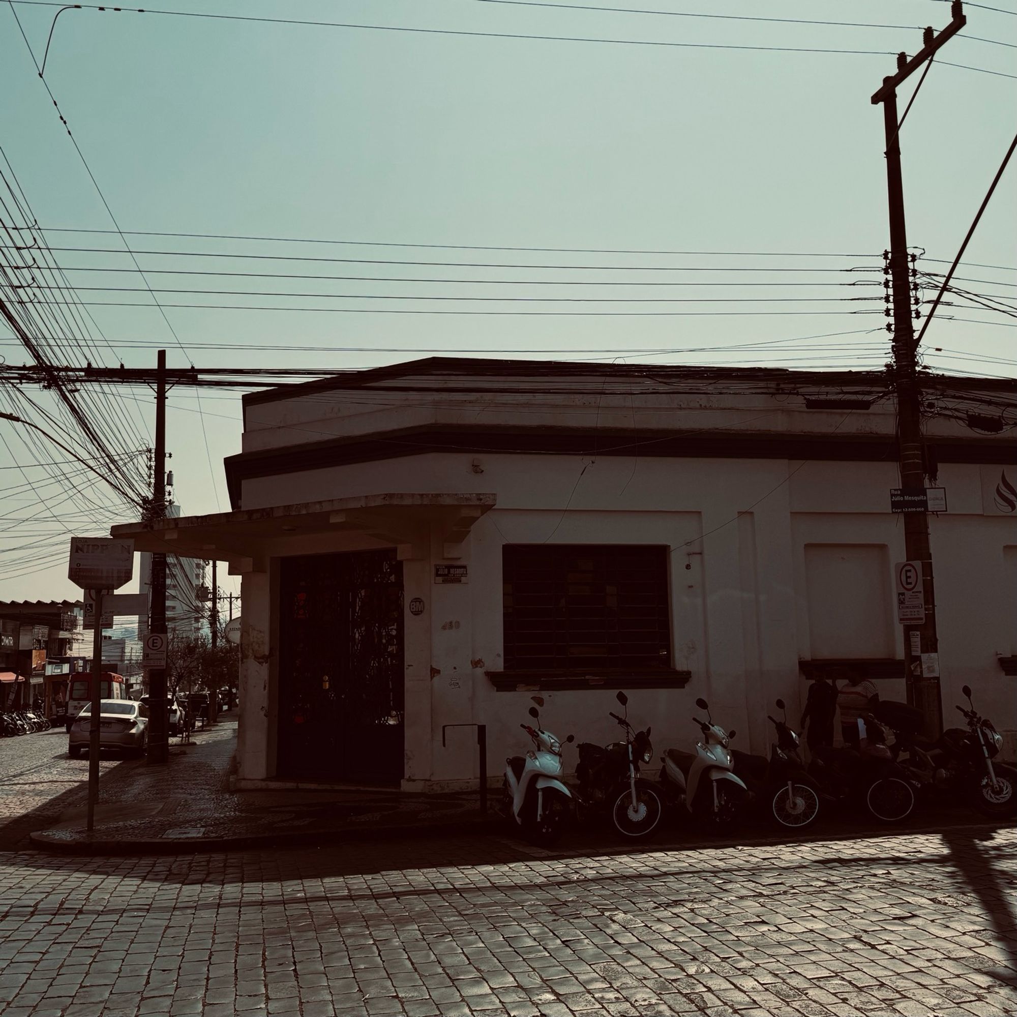 Um estabelecimento de comércio antigo, com portas fechas, numa esquina em rua de paralelepípedo. Várias motos paradas uma do lado da outra e duas pessoas andando na calçada. É possível ver os fios de energia elétrica. Foi usado um filtro quente na fotografia.