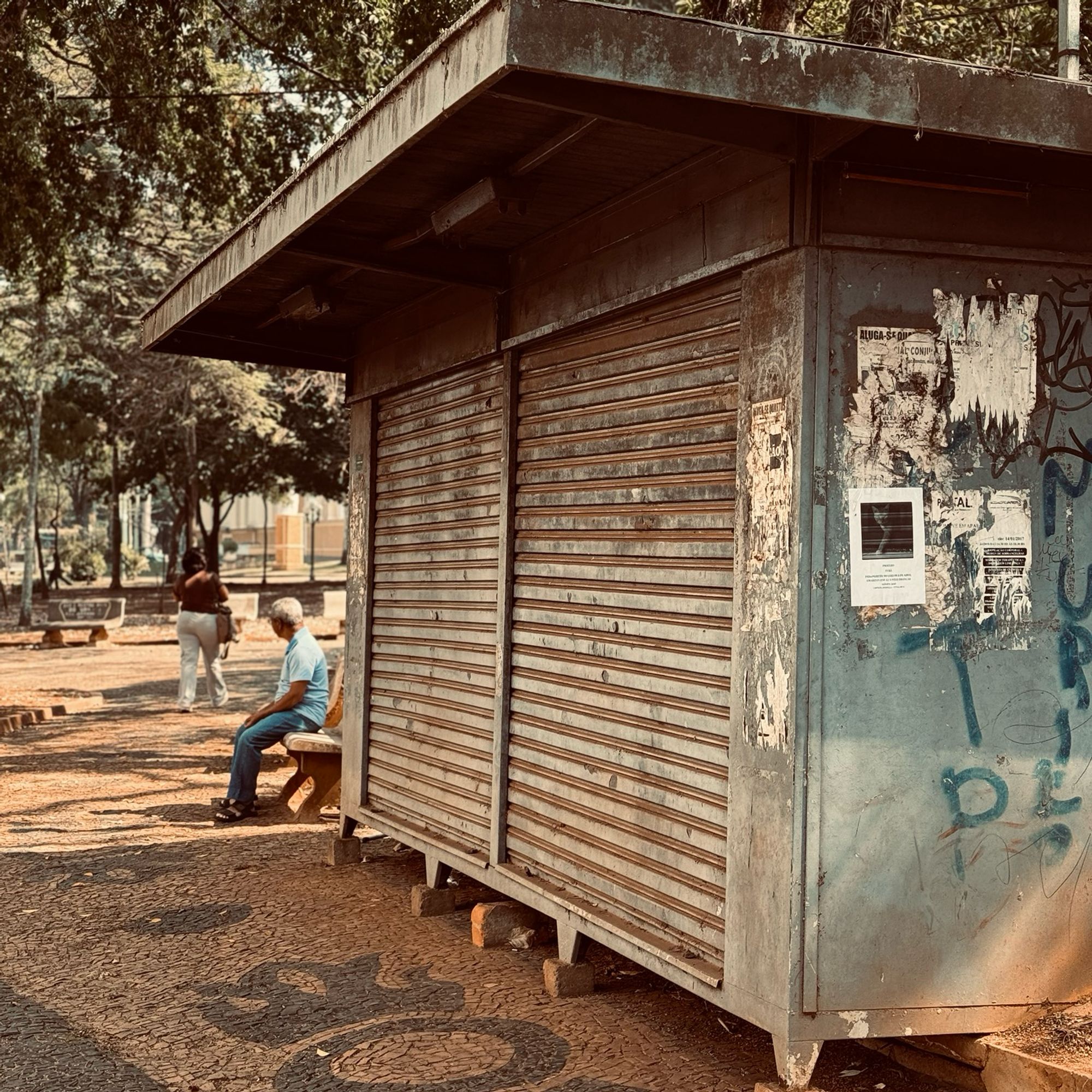 Foto com um filtro de cores quentes de uma banca de jornal com suas portas fechadas, ao fundo vemos um senhor sentado no banco da praça olhando para o lado oposto da câmera e uma senhora que parece estar caminhando também para o lado oposto da câmera.