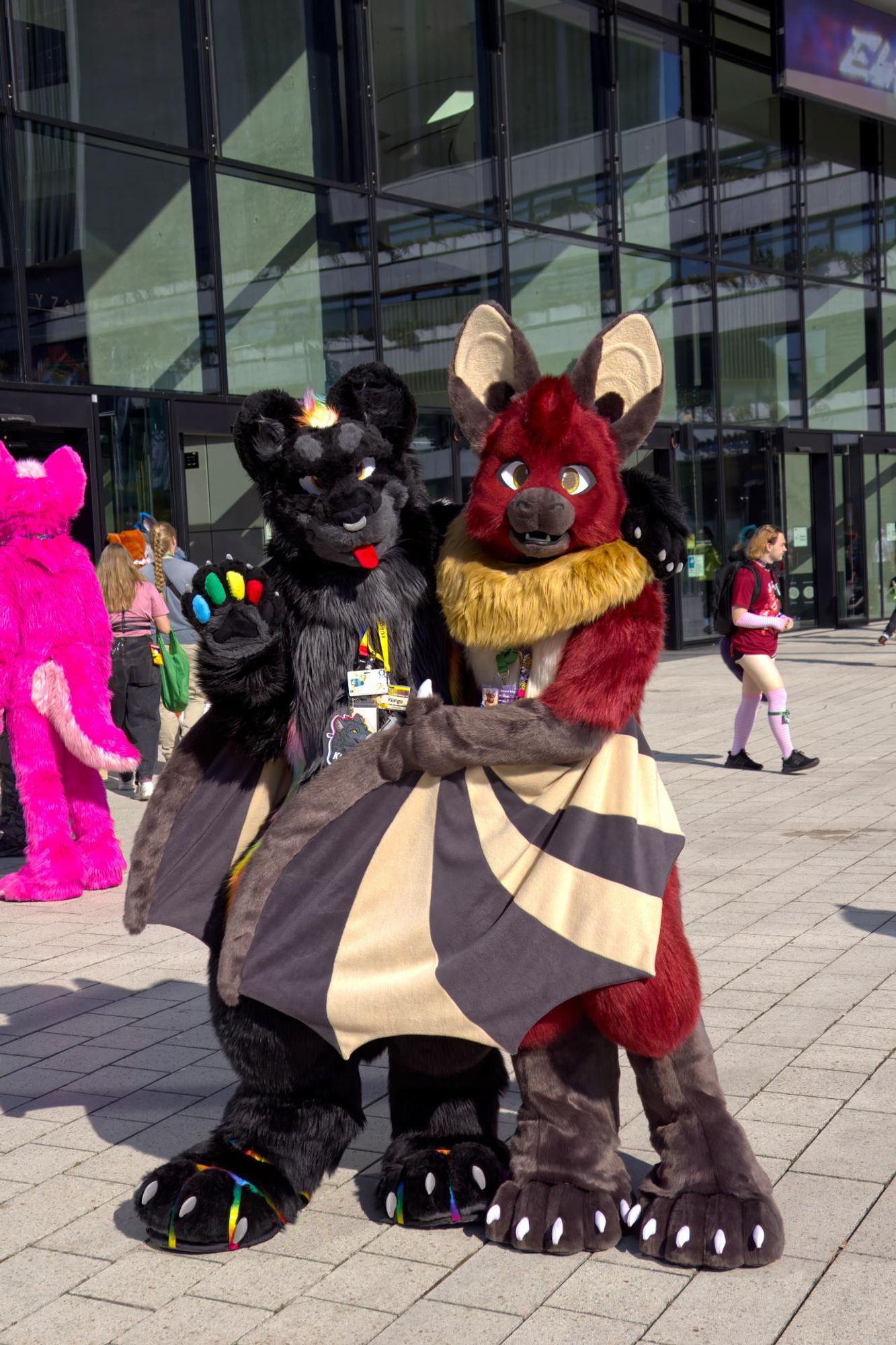 Photo of me in black jaguar fursuit, standing next to a dragon-bat fursuiter with red fur, gray paws, and beige highlights. We're both looking at the camera while I'm waving, with the bat's wings half wrapped around me.