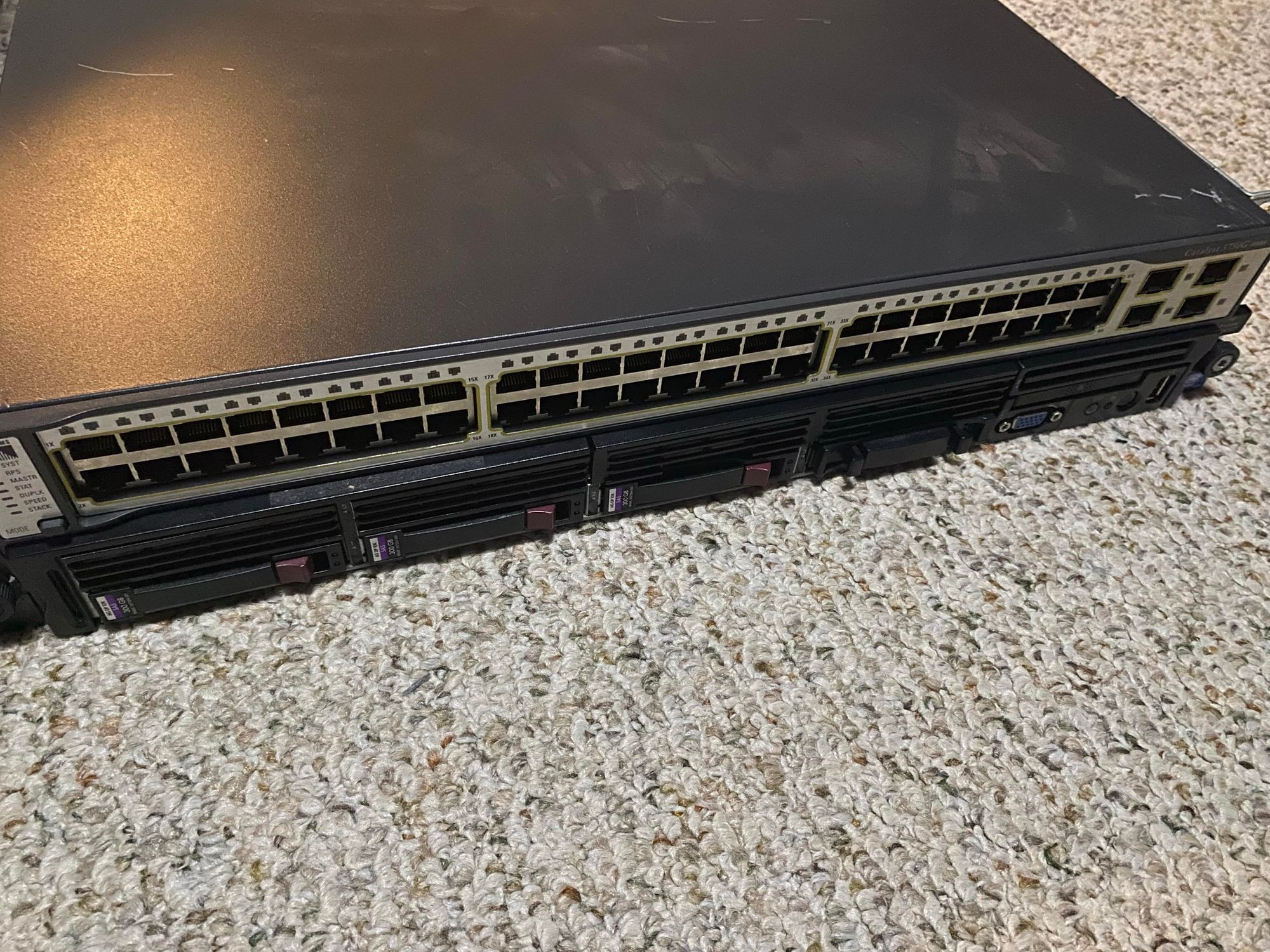 A large, dark grey 1U network switch about the size of a pizza box , covered in RJ45 network ports and a few SFP ports.  Under it is a black 1U box with 4 hard drive bays, three of which are populated with trays and the last one with a blanking plate.