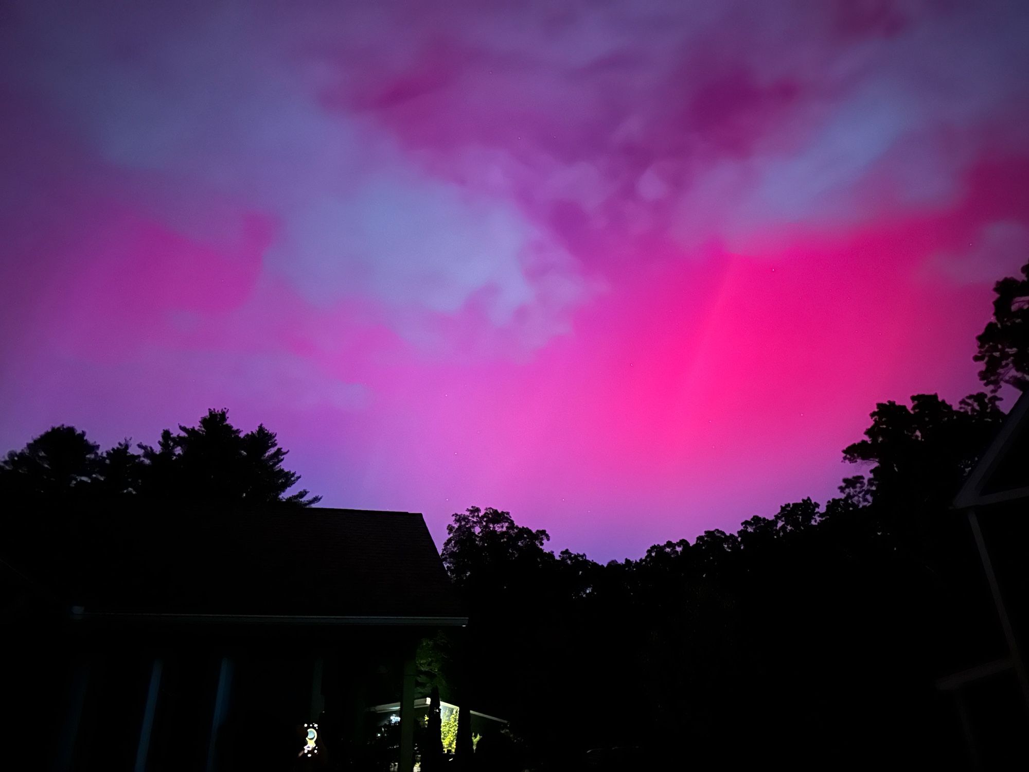 Photo of the nighttime sky over woods and houses. The Aurora Borealis can be seen as a big cloud of bright magenta.