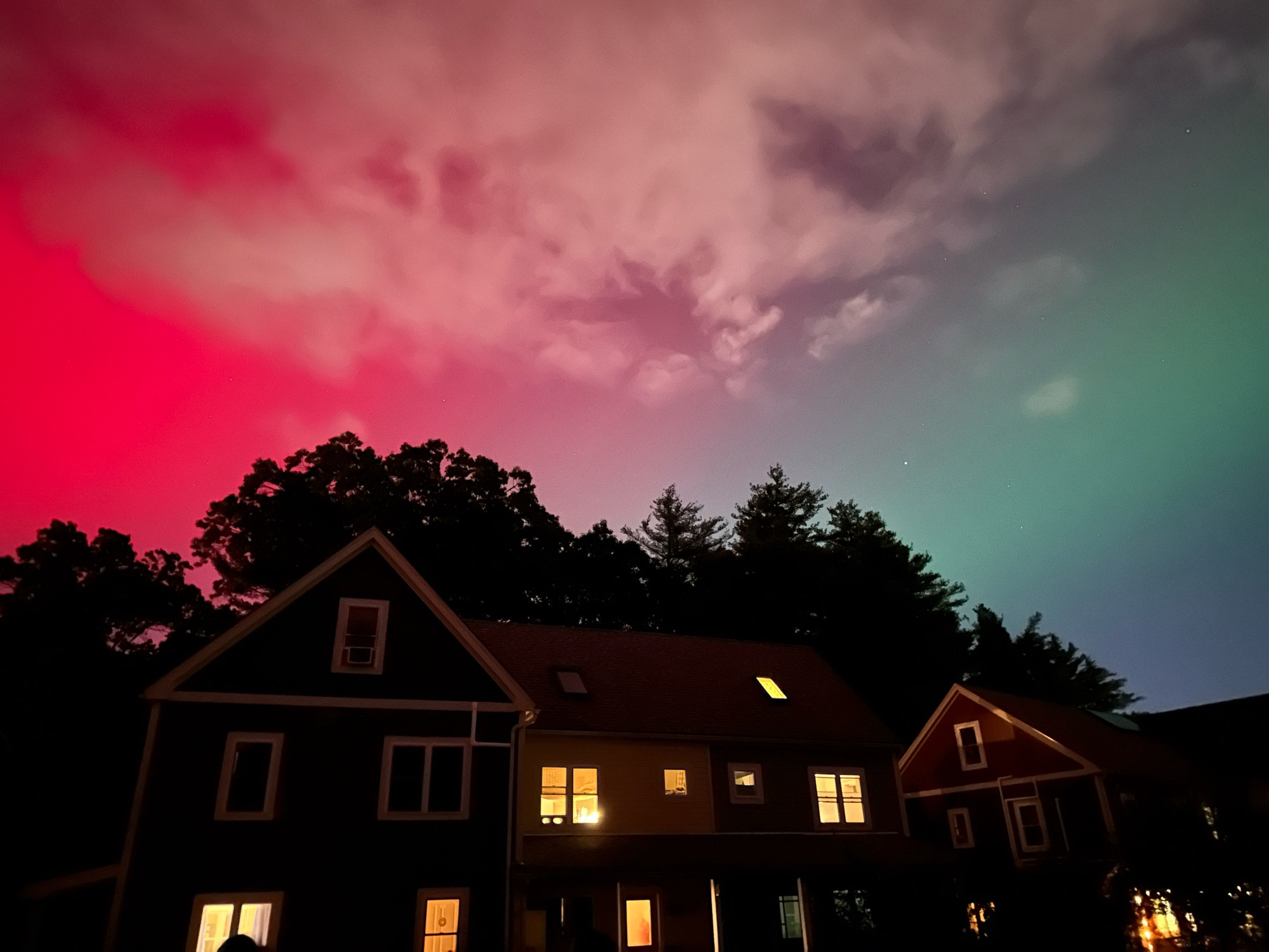 Photo of the nighttime sky over a three-story house with trees behind it. The Aurora Borealis can be seen with waves of bright magenta red on the left and soft teal green on the right.