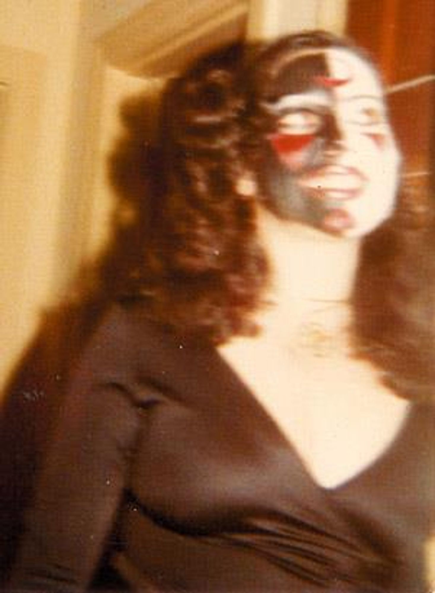 Claudia Canuta de Menezes, a woman in a low cut black top and thick curly black hair.  She has large eyes  She is wearing face paint.  One side is painted white, the other black.  Various red highlights on the eyebrow, below the eyes, and on the chin are present.