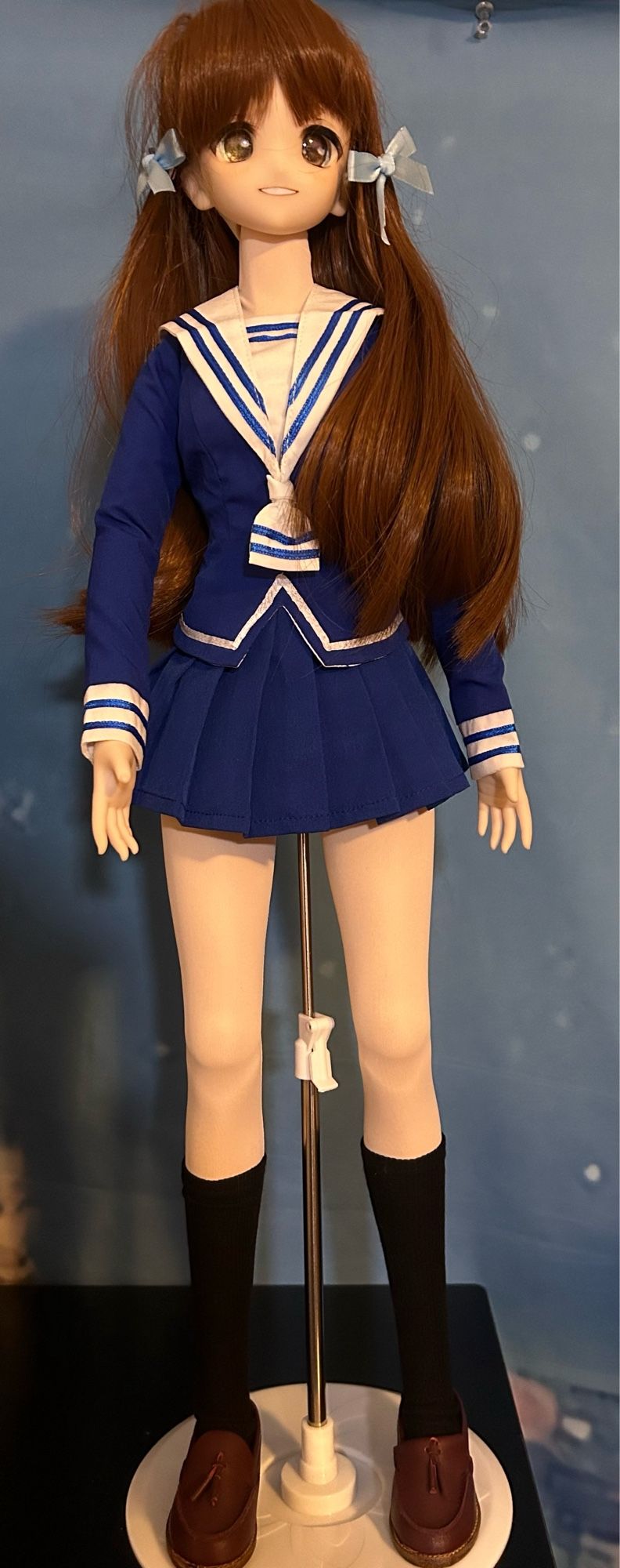 A Dollfie Dream Sister with brown hair and eyes, dressed in the school uniform of Tohru Honda from Fruits Basket, which is a blue school uniform with a white collar, cuffs and trim, a blue skirt, black socks and brown loafers. She has blue ribbons in her long brown hair.