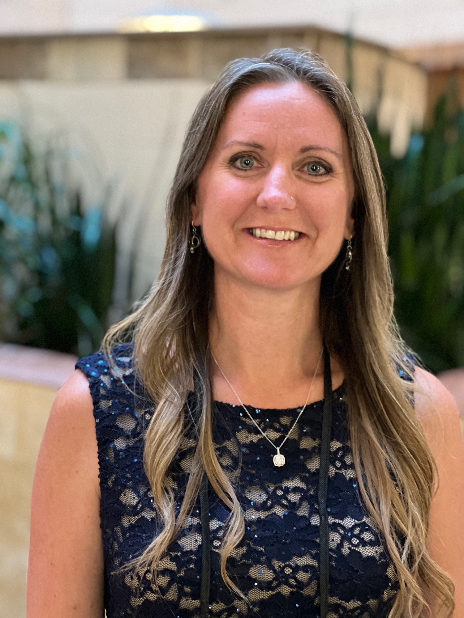 Kat, a white woman with blue eyes, long wavy dark blonde hair, smiling with teeth, wearing a navy blue sleeveless knee-length dress that’s solid and scalloped from the waist down and lacy from the waist up. She’s standing in a hotel lobby about to attend an awards dinner where she gave a speech on never giving up as writers!