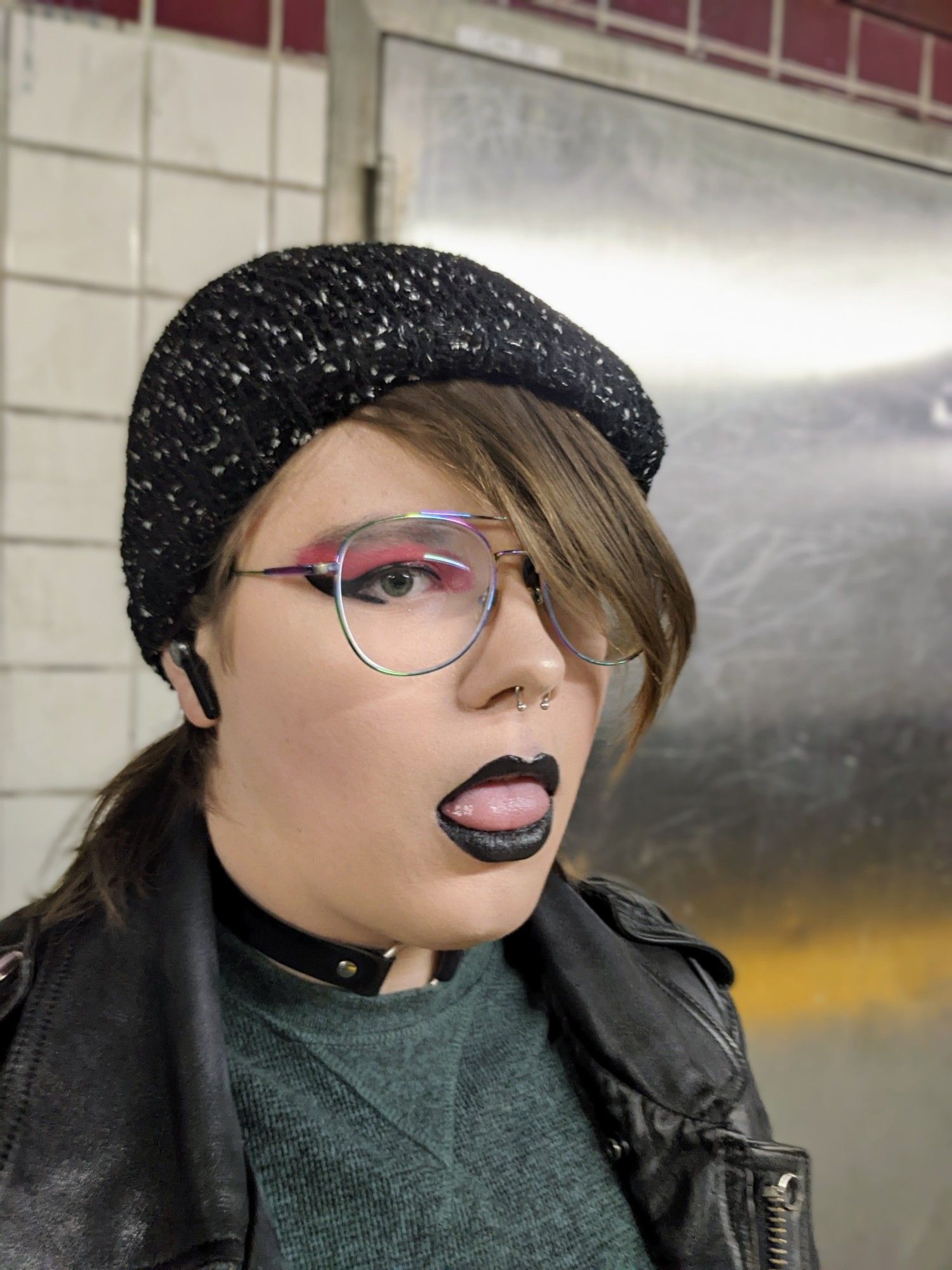 Percy selfie in the septa underground, she's wearing her leather jacket and a green shirt. she's got a chocker and her gf's beanie on. she's sticking out her tongue and has black lip stick on