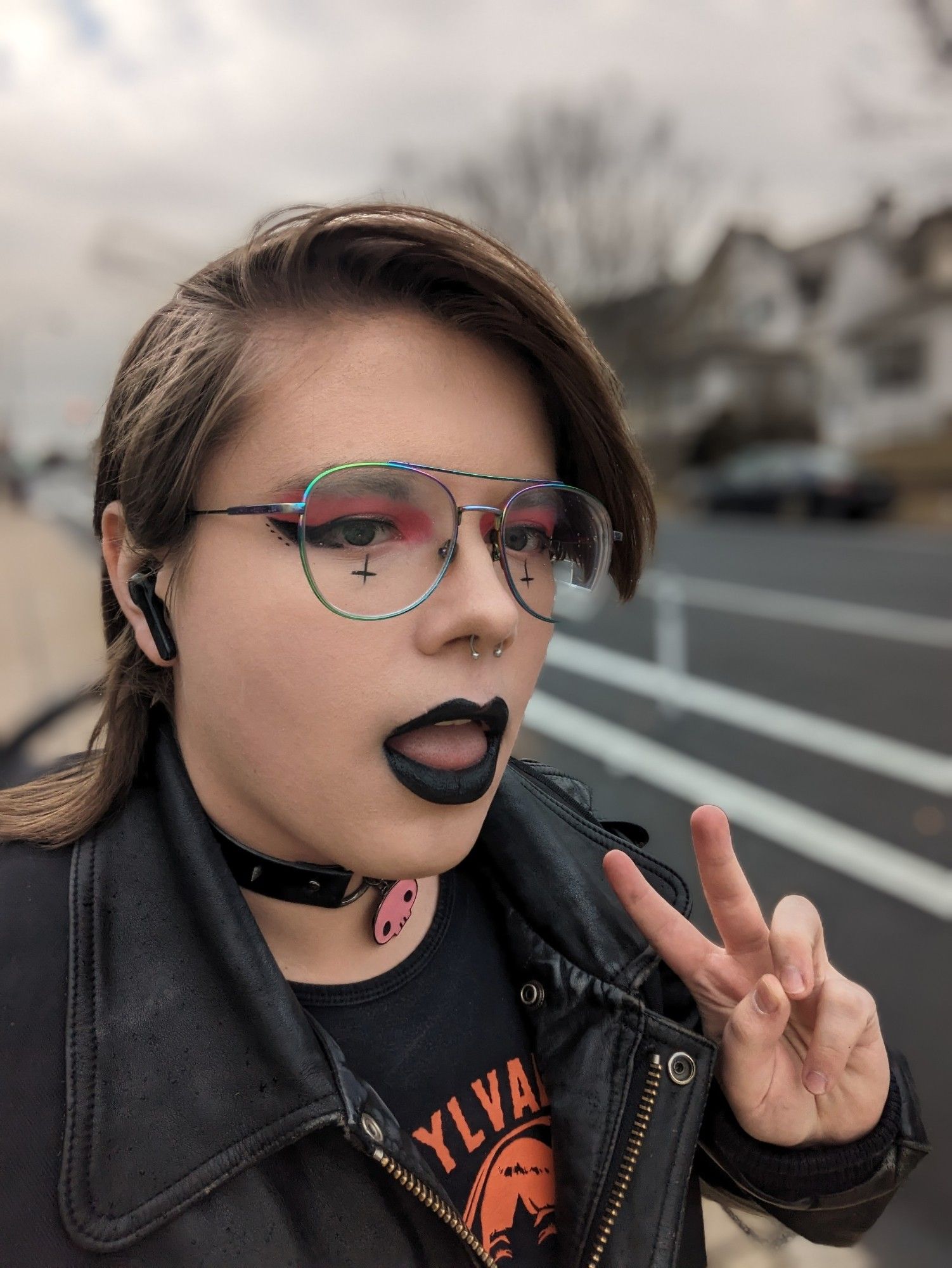 Outdoor selfie of Percy. She's wearing a black T-shirt with an orange graphic design, her leather jacket, a spiked collar, and full foundation makeup. Her eyeshadow is a sparkly reddish-pink and she's drawn upside down crosses with eyeliner under each of her eyes. She has her left hand left up towards the camera with a peace sign ✌️