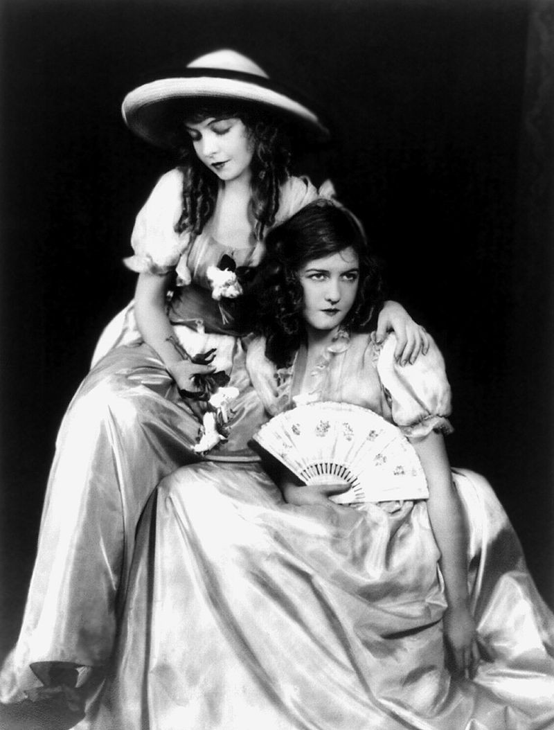 Dorothy and Lillian Gish in publicity photograph for film, 1921. Black and white. Lillian is sitting down a little above Dorothy. Lillian is wearing a hat and she has long, curled, dark hair. Both Lillian and Dorothy are wearing long dresses, with short sleeves. Dorothy is sitting down a little below her sister, she is holding a fan. Her hair is dark, curly and medium sized.