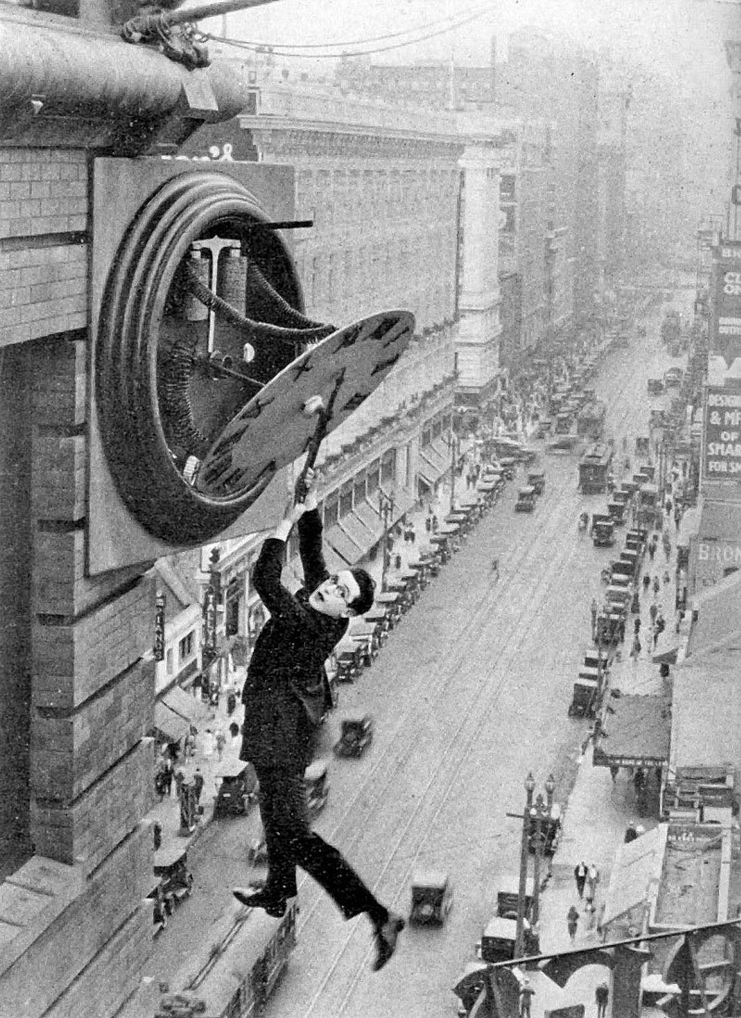 Harold Lloys is hanging from a clock. He is wearing his trademark glasses, black suit and shoes. We can see buildings around him and the street with cars below him.