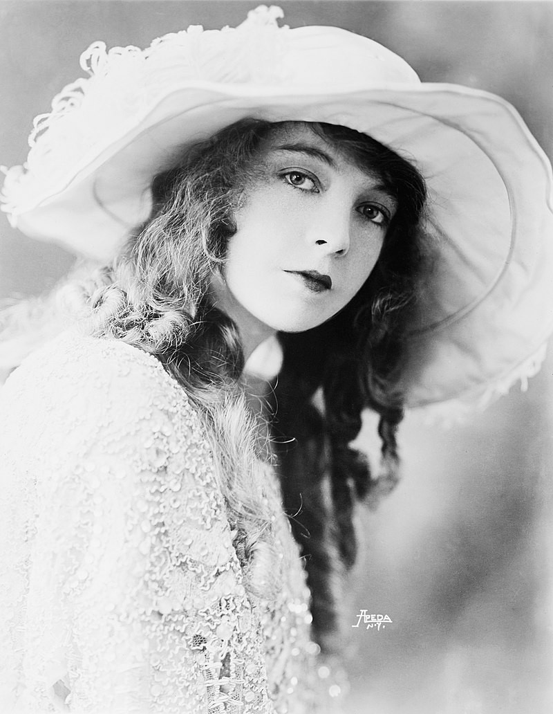 Lillian Gish. 1921 publicity photo. Black and white. Her hair is down, long, with curls. She is wearing a hat and a dress, both of them in light colors. 