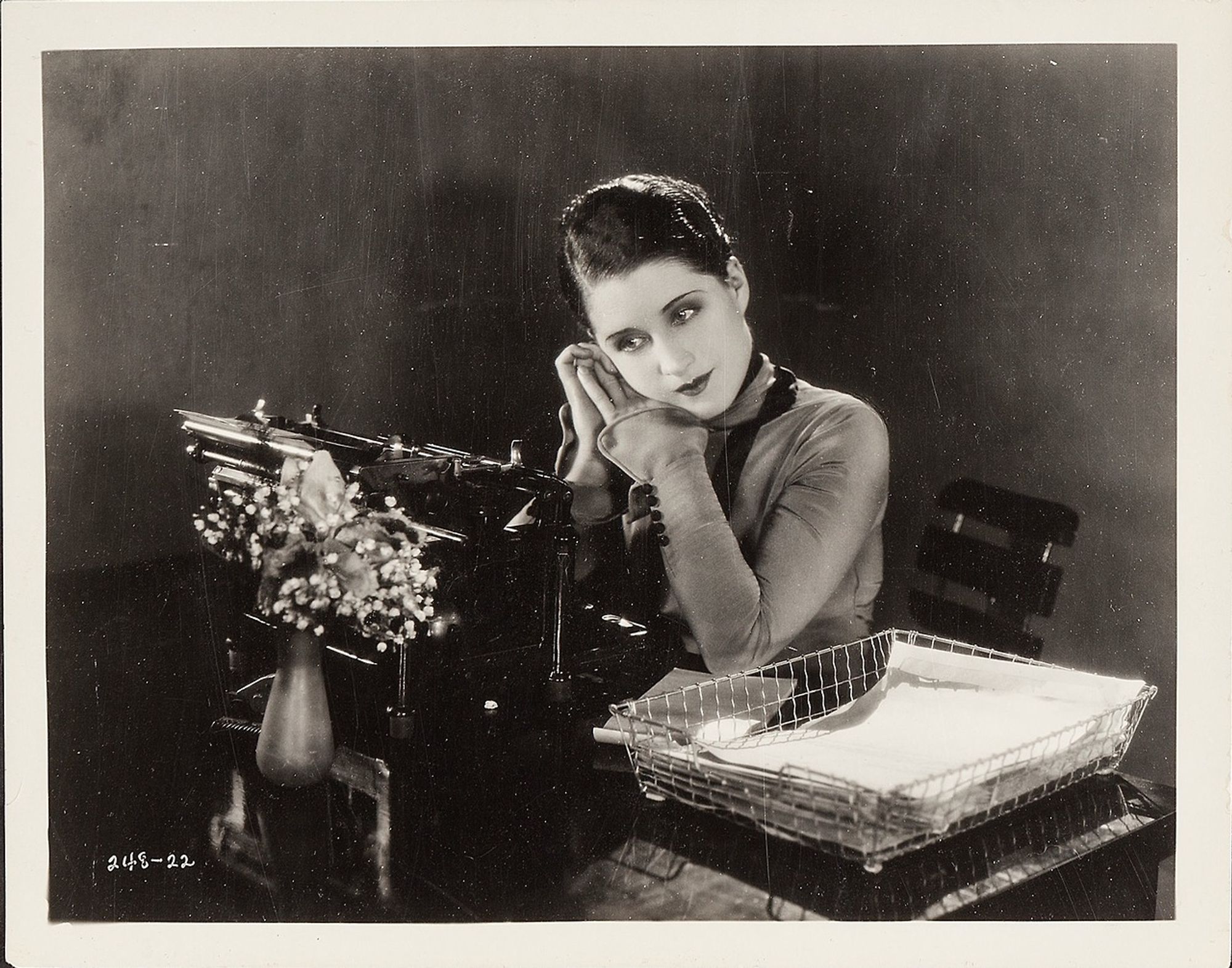 A woman is sitting behind a desk. We can see on the desk a typewriter, a vase of flowers and some papers. The woman is wearing a long-sleeved dress. Her hair is dark and wavy. She has both elbows on the desk. 