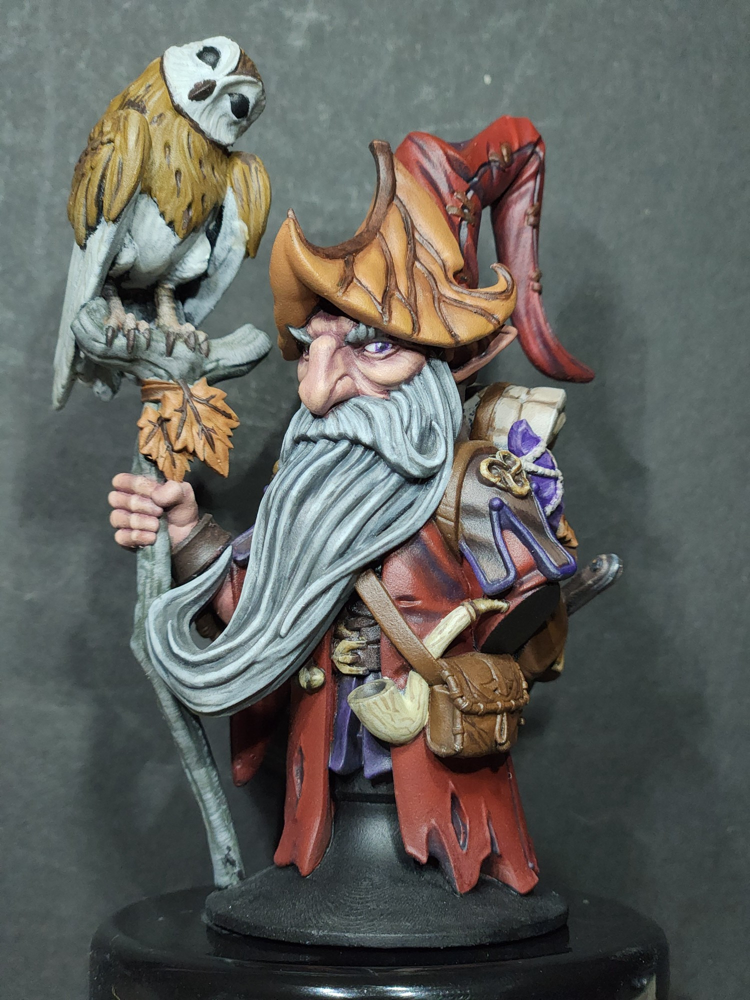 A bust of a gnome with a red cloak and pointy hat holding a walking stick with an owl perched on top.